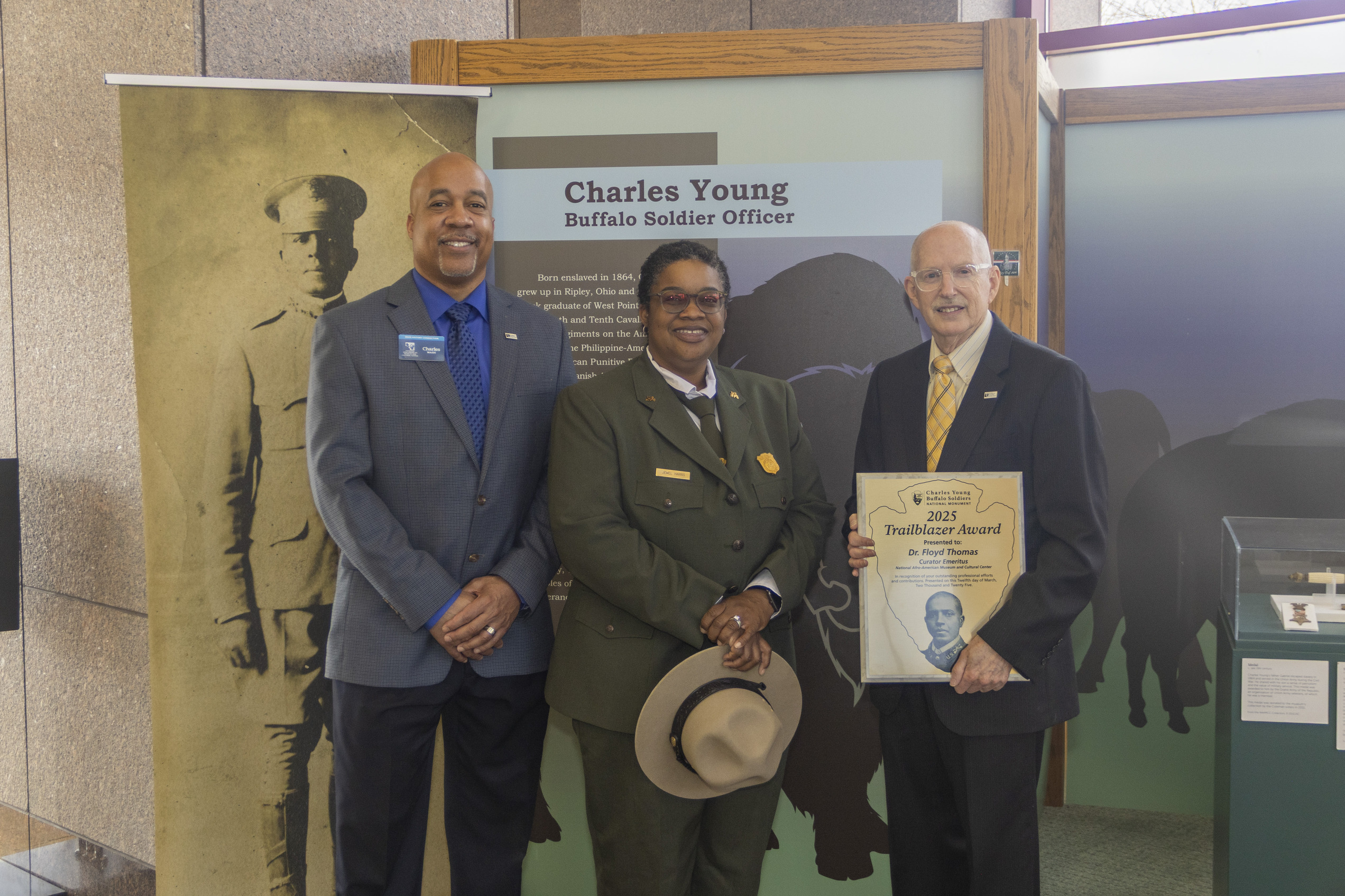A man wearing a blue-colored suit stands to the left, a park ranger in a green uniform holding a tan flat hat stands to his left, a man wearing a dark-colored suit stands to the far right.