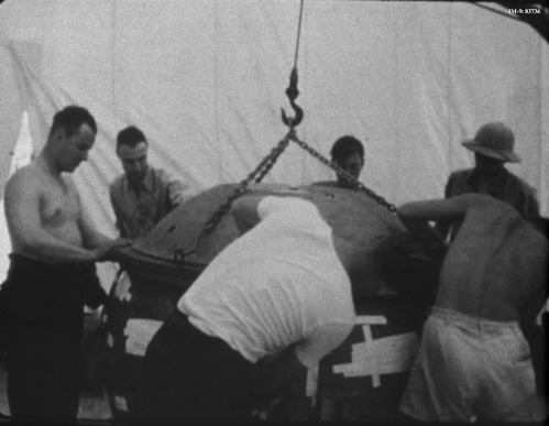Black and white photo of six men around a large metal sphere attached by chains to a hook. They are leaning over, checking its security. Some men are shirtless, others in work clothes. 