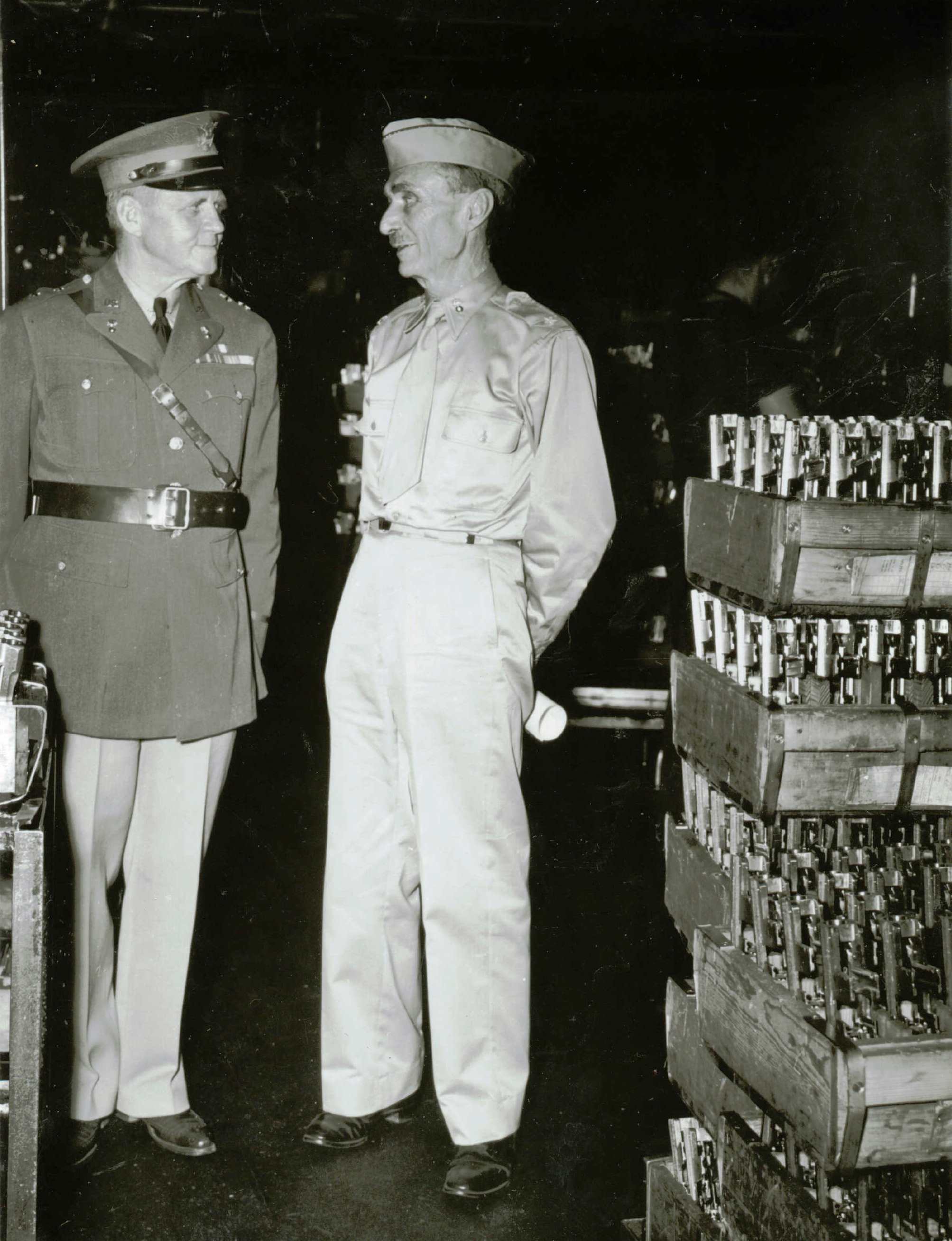 Black and white photograph of two men in military uniform standing by crates of metal objects.
