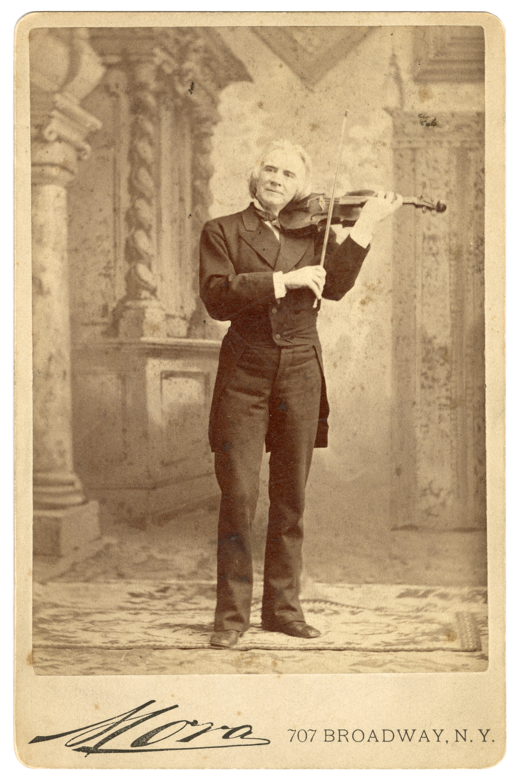 Full-length portrait of a white man playing violin in dark formal attire. The backdrop is decorated with twisted columns and a curtain. Man stands on a carpet on top of another carpet.