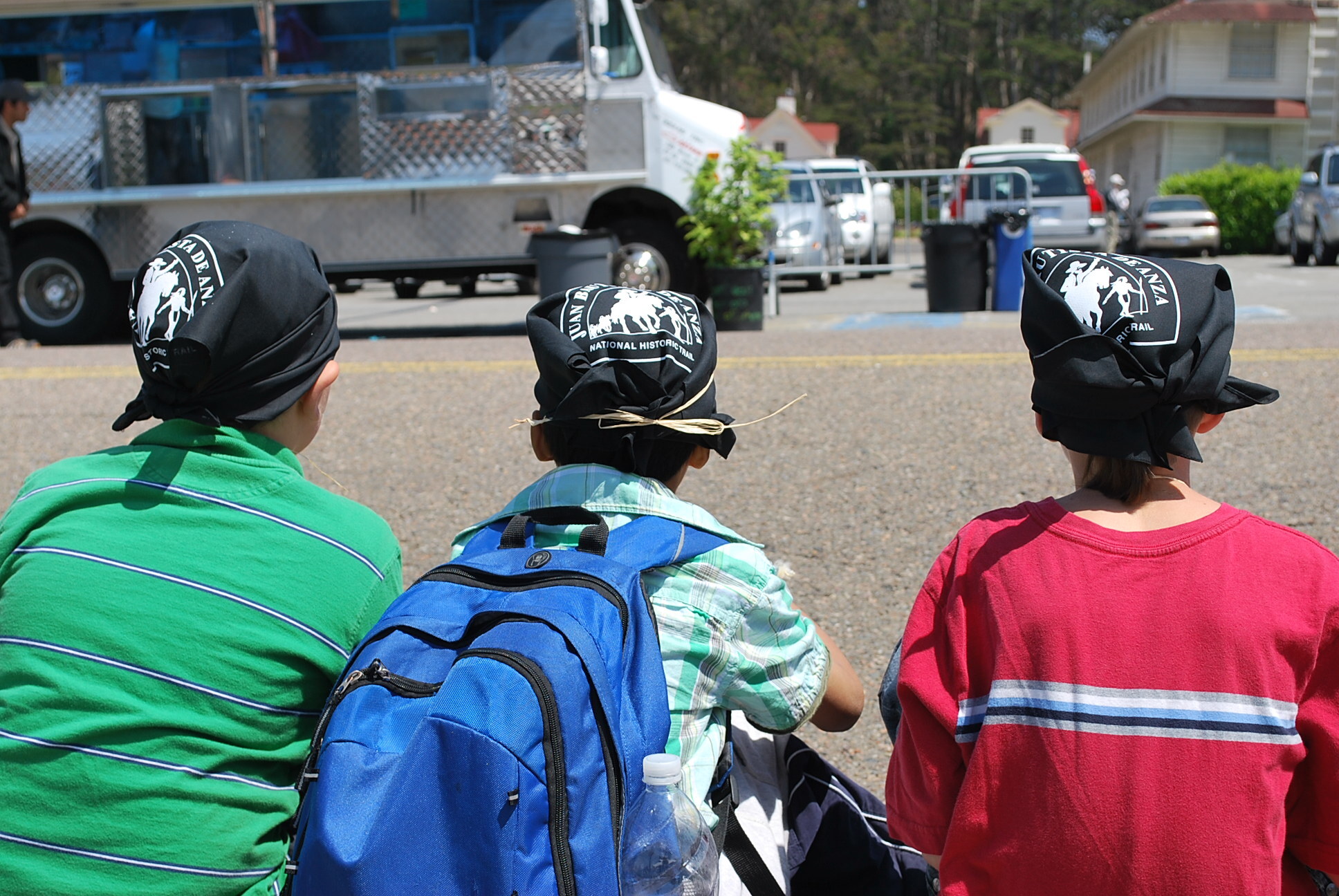 Three kids sit on a curb wearing black Anza Trail bandanas on their heads
