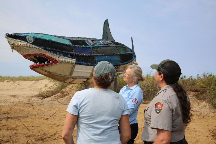 Three women, one in National Park Service Uniform" look at a 14-foot sculpture of a white shark made out of marine debris attached to a metal frame. The sculpture is located on a sandy beach area.