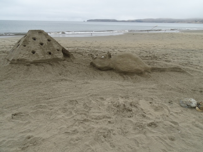 A medium-sized sand sculpture of mouse facing a pyramidal-shaped block of Swiss cheese.