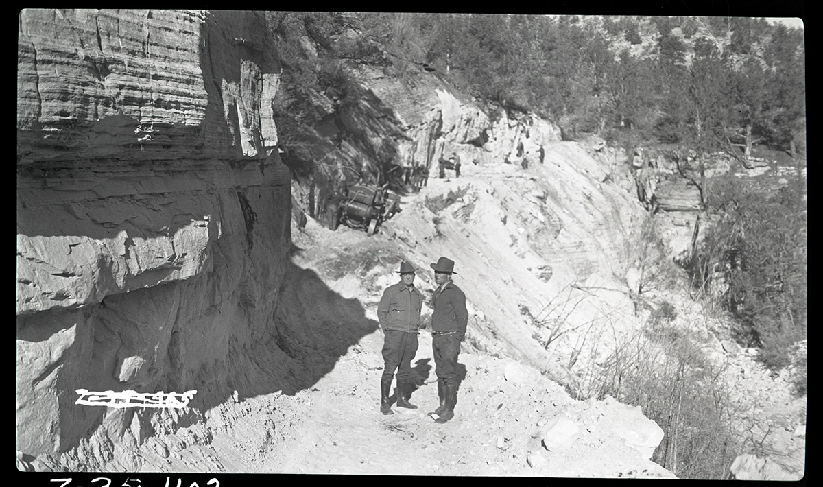 Two men, on right: Chief Ranger Donal Jolley Standing on East Rim Trail, then under construction, along edge of white cliffs. Civilian Conservation Corps (CCC) work crew and equipment in background.