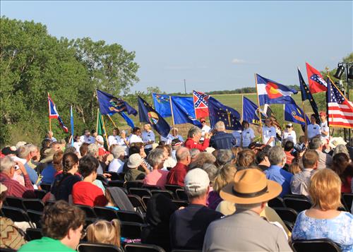 A crowd of people sitting outside. Many flags from various states are at the front of the group.