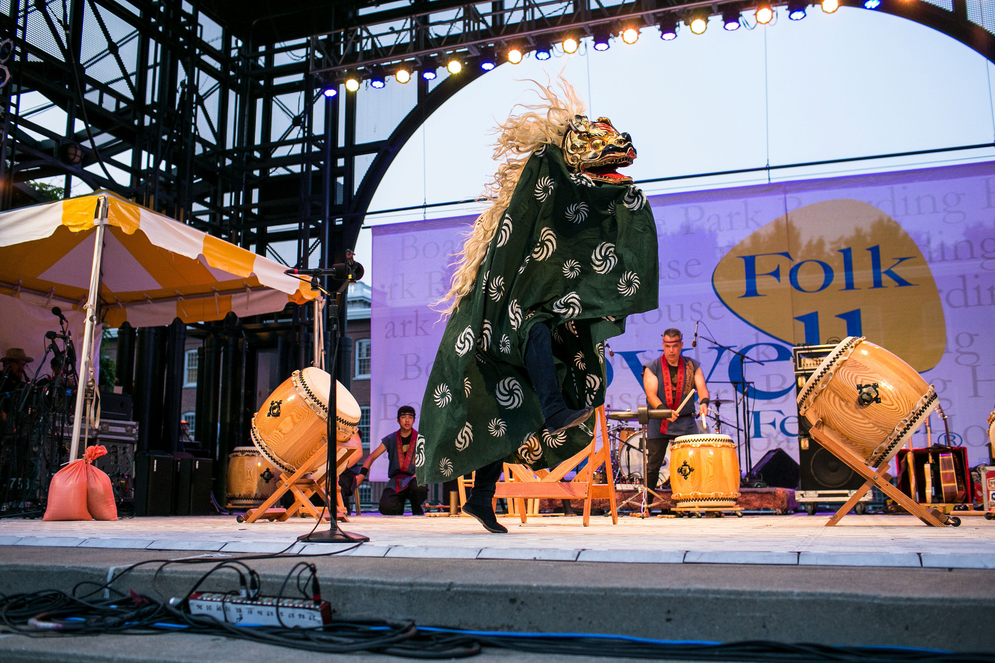 A performer, on stage, is dressed in a lion costume, with one foot elevated and the other planted to spring upward. The lion has a golden face with a wide, open mouth. It has a long mane of flaxen hair, and its body is green fabric with white concentric swirls. There are taiko drummers in the background.