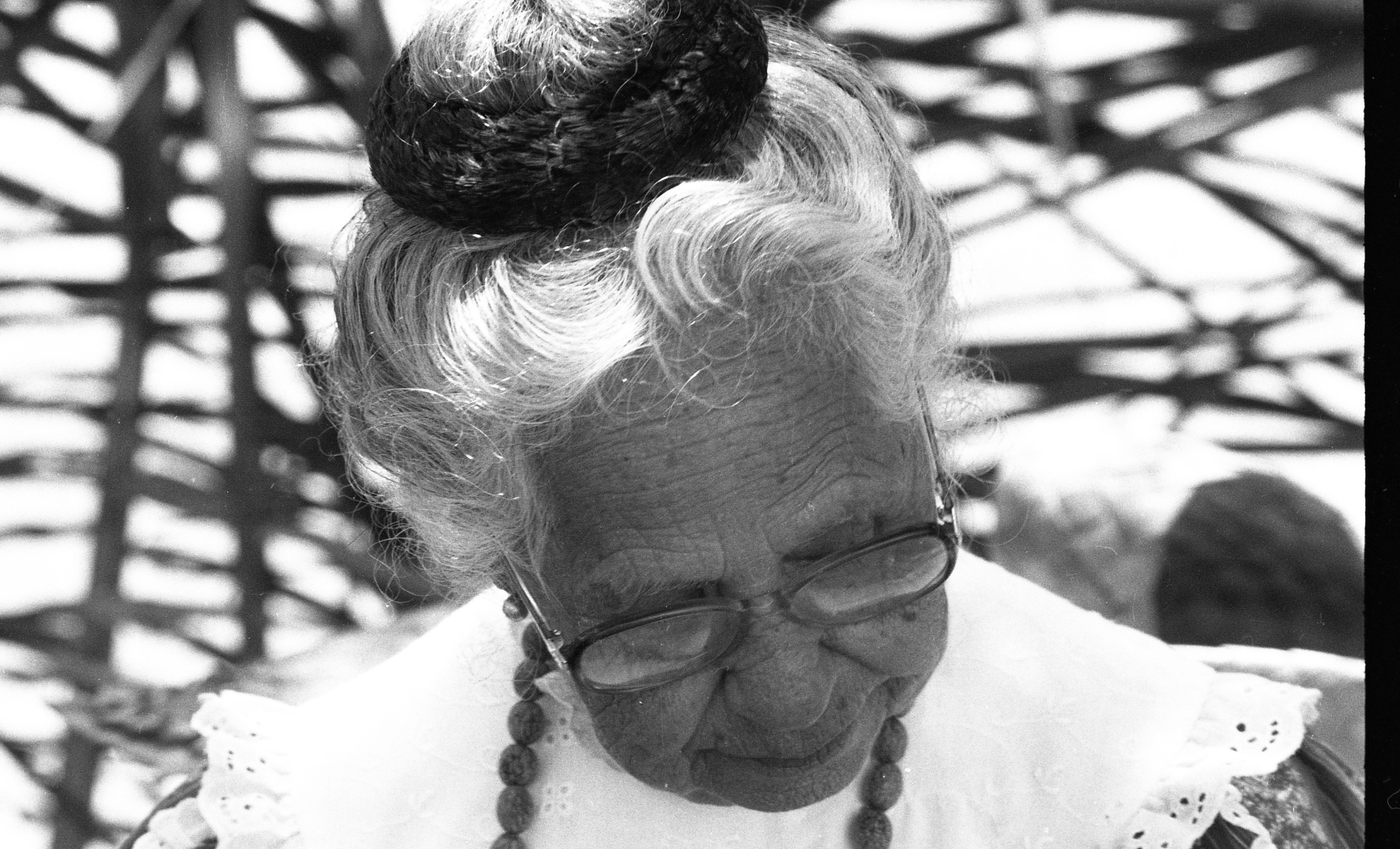 A black and white close-up head shot image of an older woman.  The older woman has her gray hair up in a ballerina bun with a thick scrunchie.  She is wearing glasses and small ball-shaped earrings. She is looking downward and not at the camera. She is also wearing a beaded necklace and a high neck Vandyke eyelet collar with a scalloped hem.