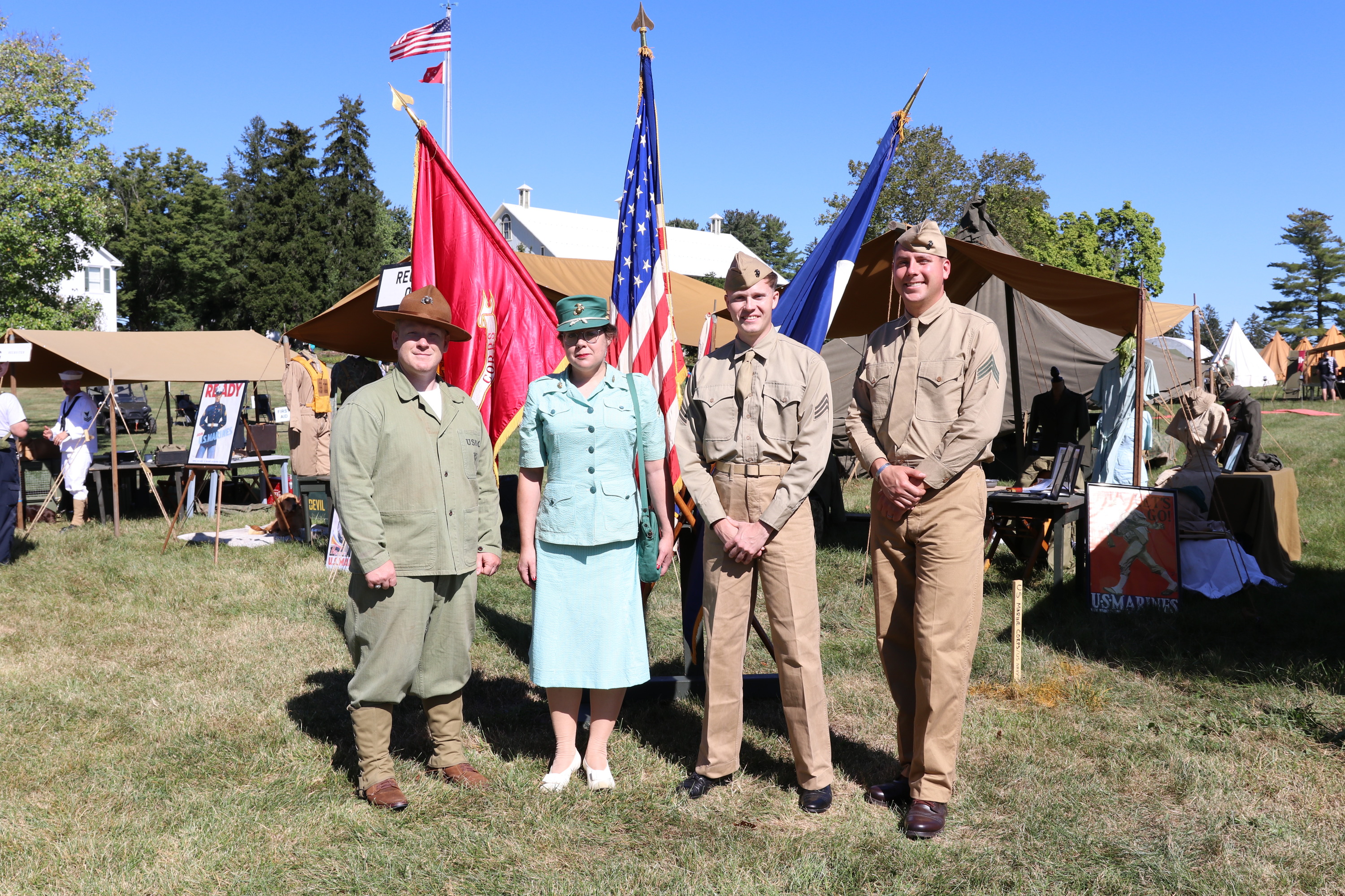 Several volunteers in historic uniforms stand in front of several flags and green military tents. 