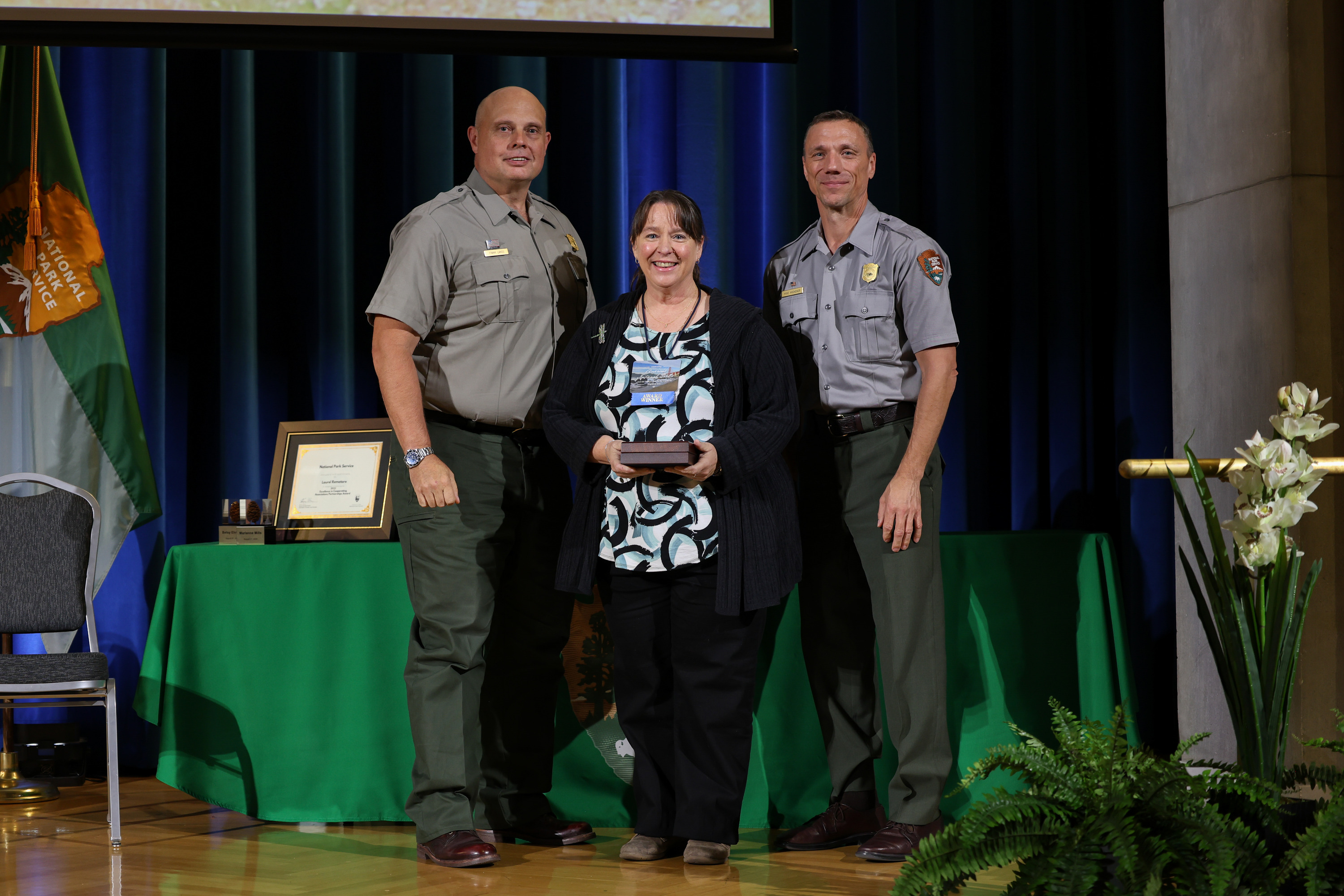A woman holds an award with two men in gray and green uniforms at an award ceremony.