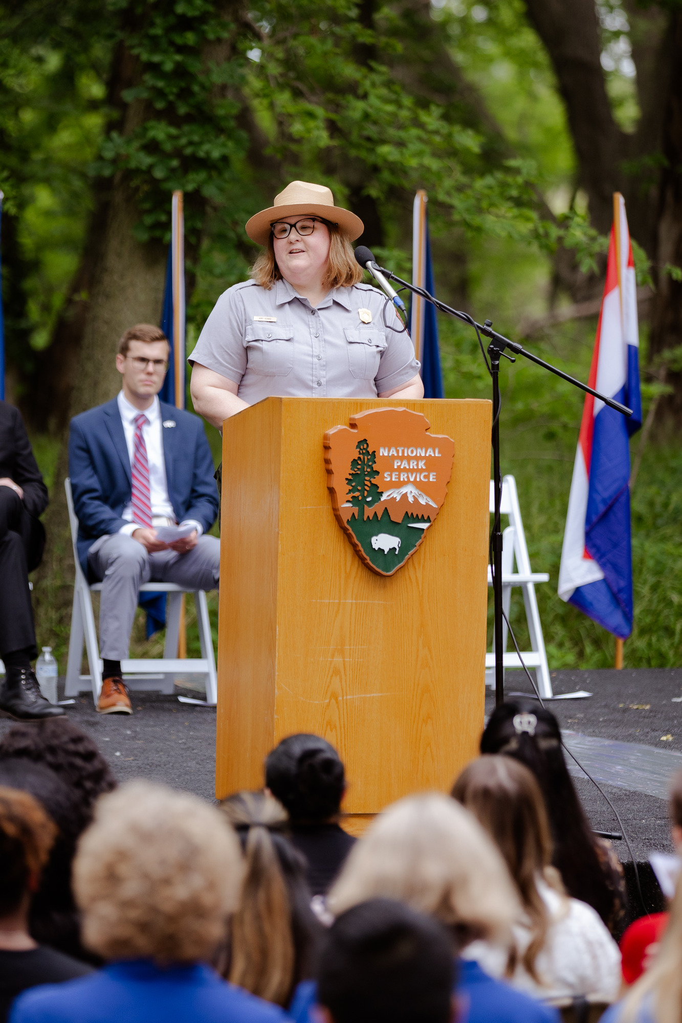 A uniformed female ranger stands at an outdoor podium with an NPS logo and looks out to the gathered audience.