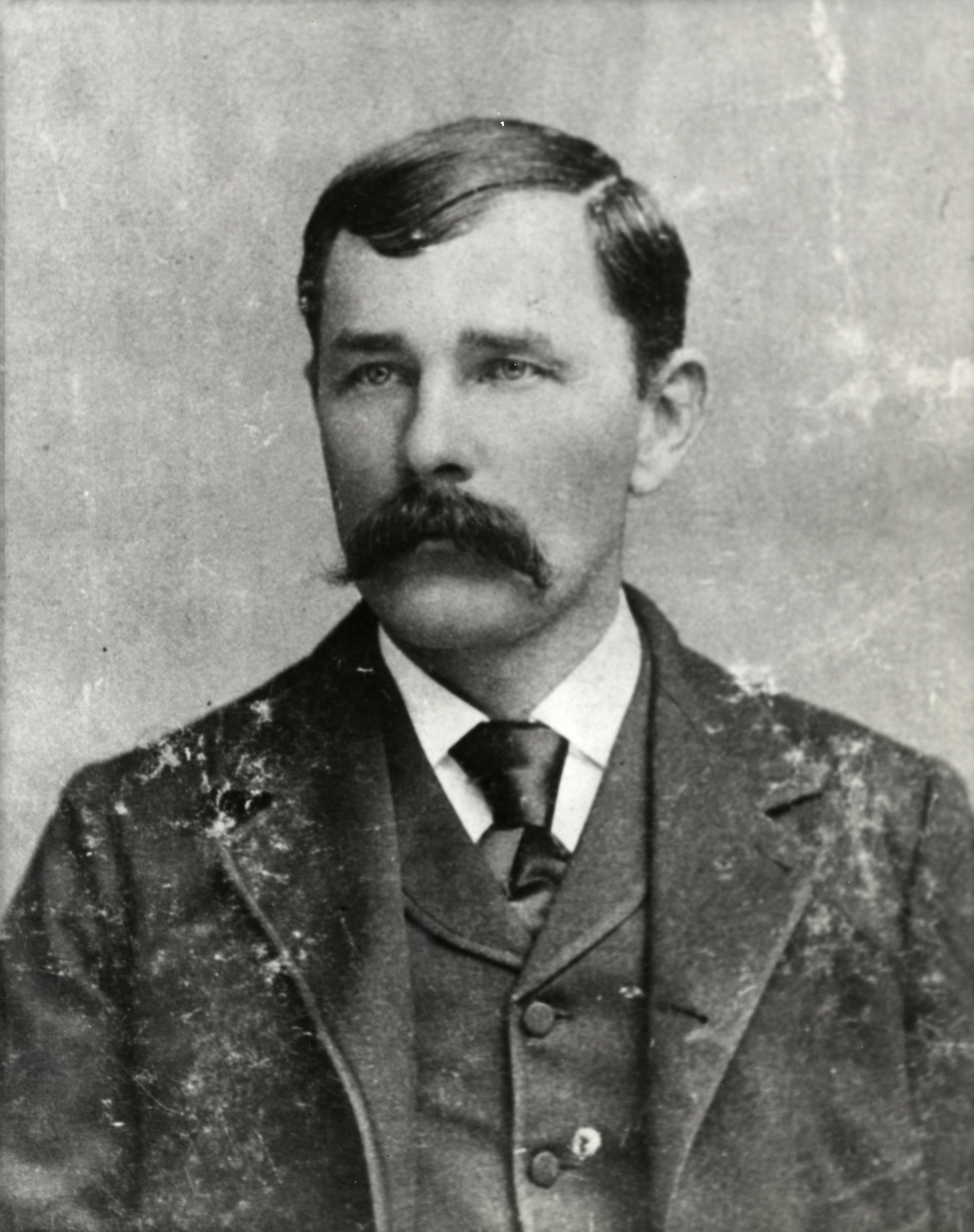 A white man with a thick mustache wearing a dark three piece suit and necktie.