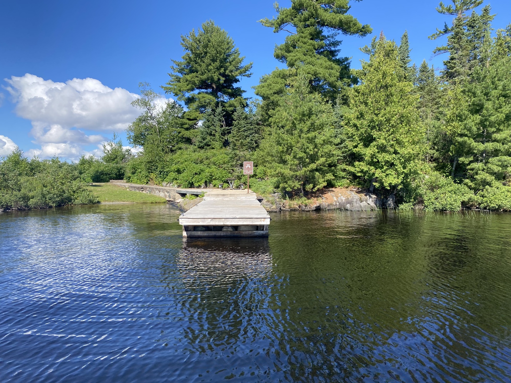 A weathered wooden dock juts into calm, rippled water, with a sign at the end and a forested shoreline under a blue sky.