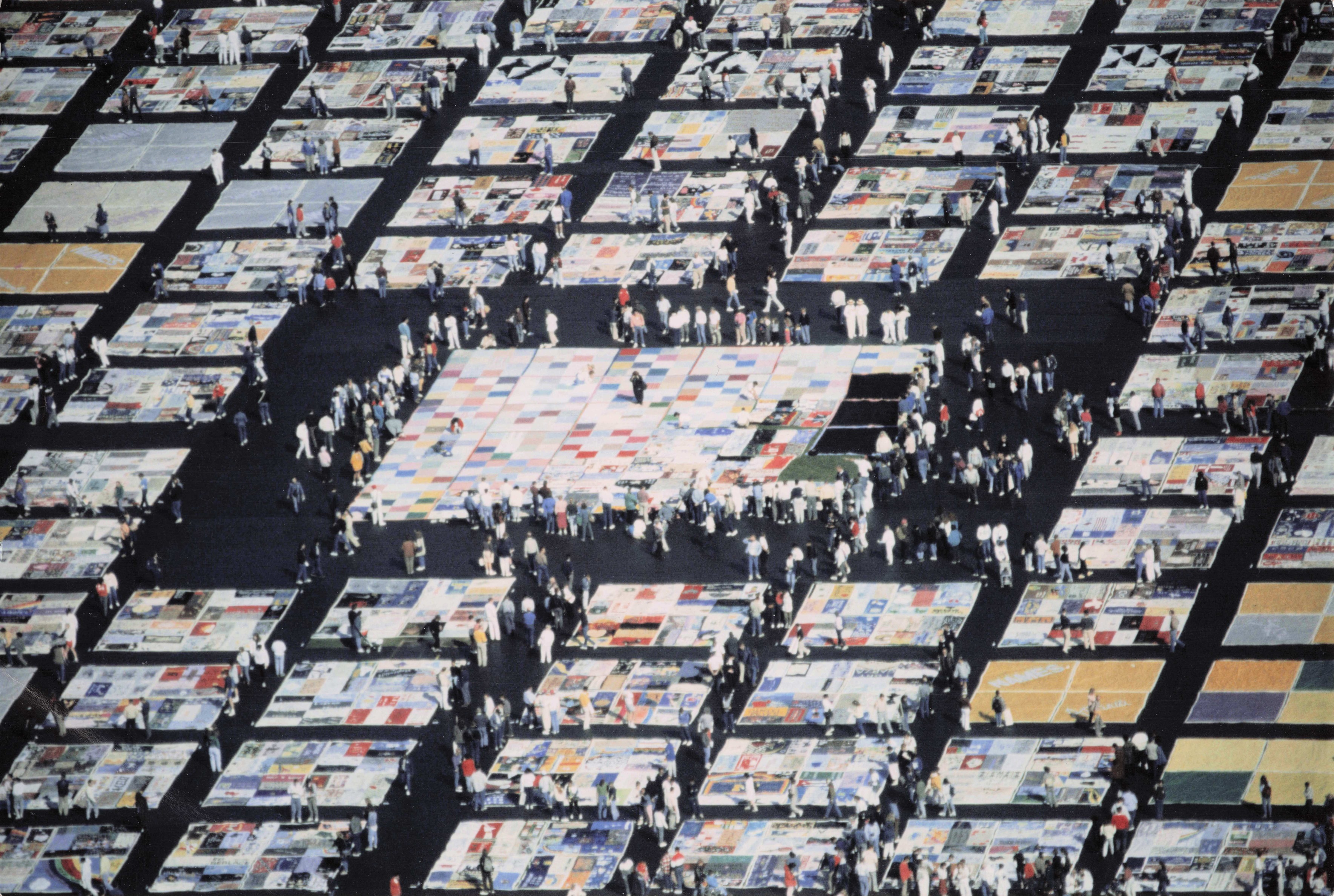 Multi-colored panels are laid on the ground. Dozens of people walk on top of the panels. 