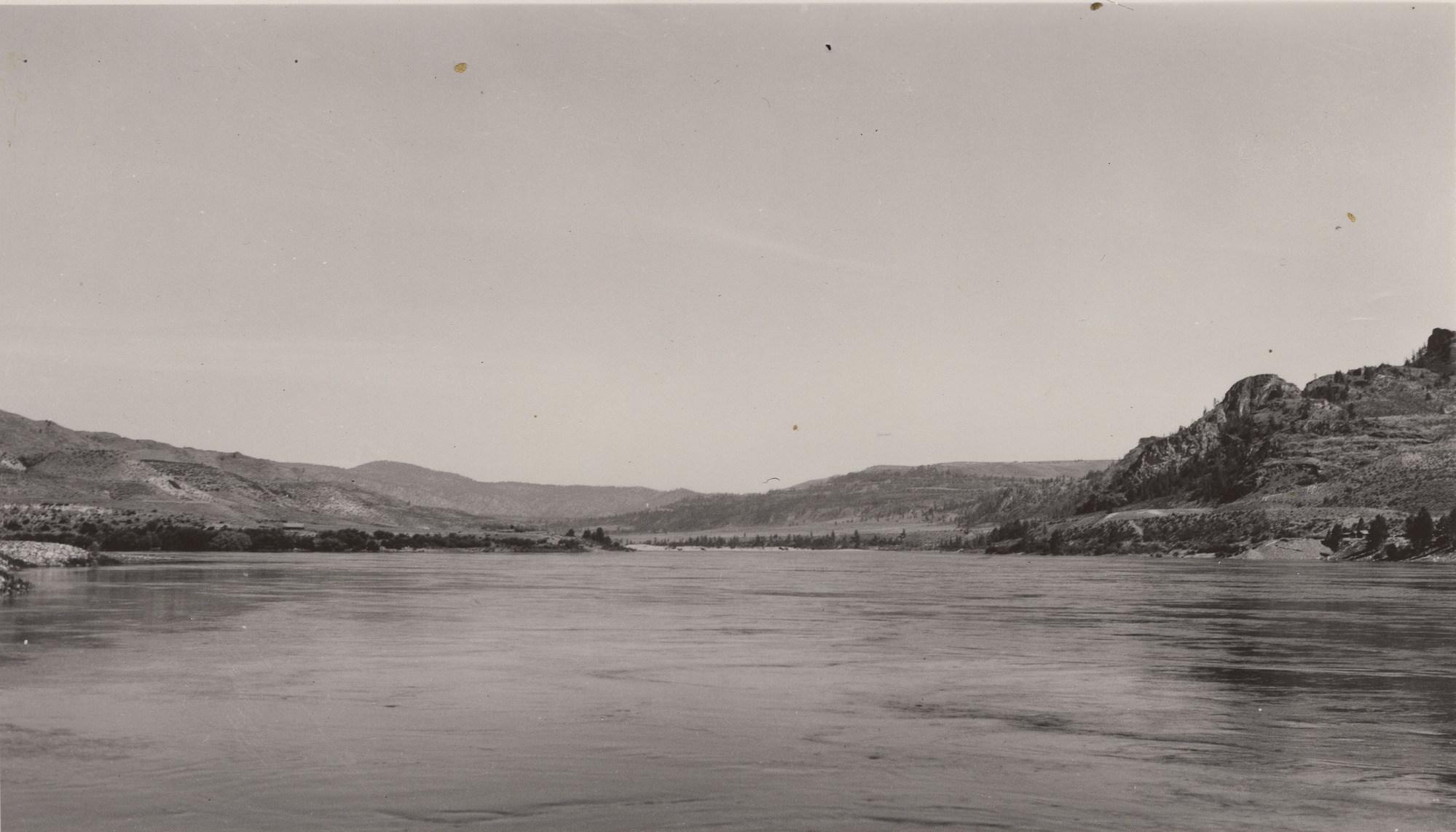 Black and white photograph taken looking across a large body of water, with rocky cliffs and gentle rolling hills in the distance