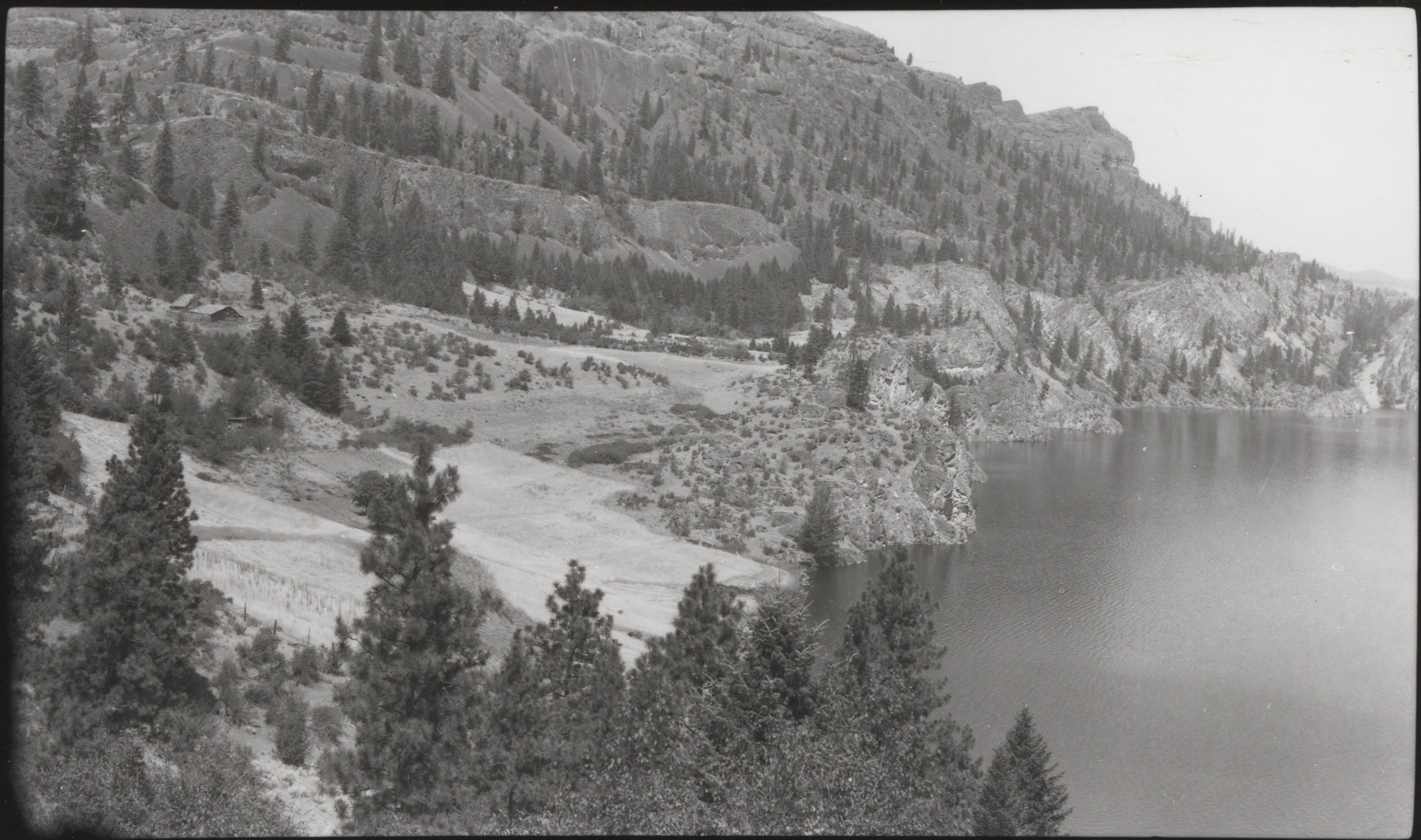 Black and white photograph of steep cliffs dotted with trees rising above a body of water.