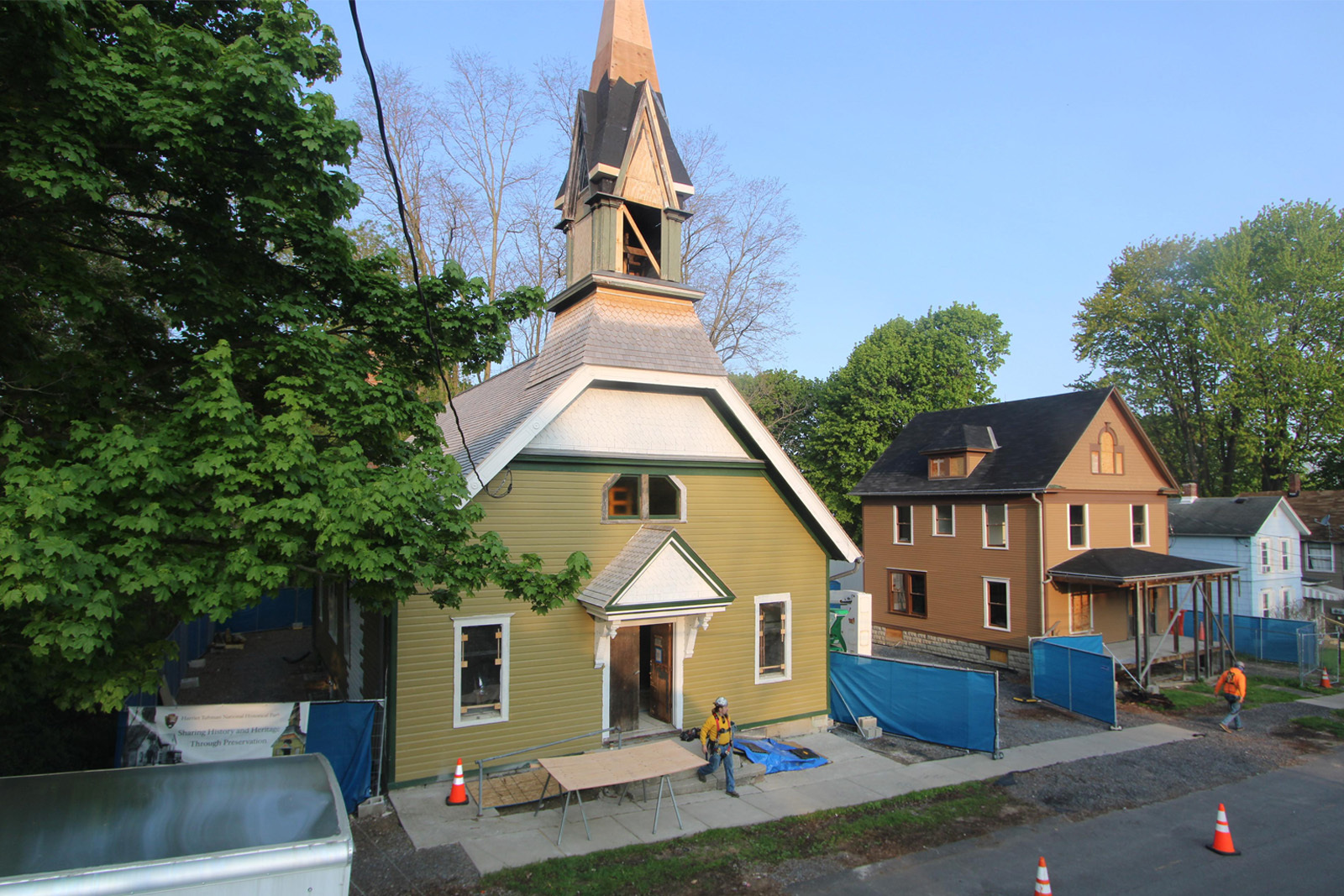 An olive-green church building with a tall unfinished steeple, to the right there is a brown house.