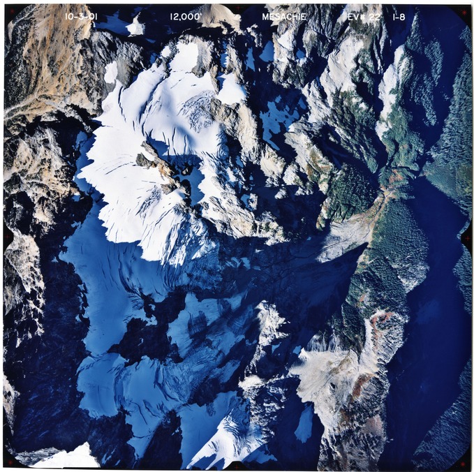 Color aerial photograph of glaciated mountain peak with forested valleys below.
