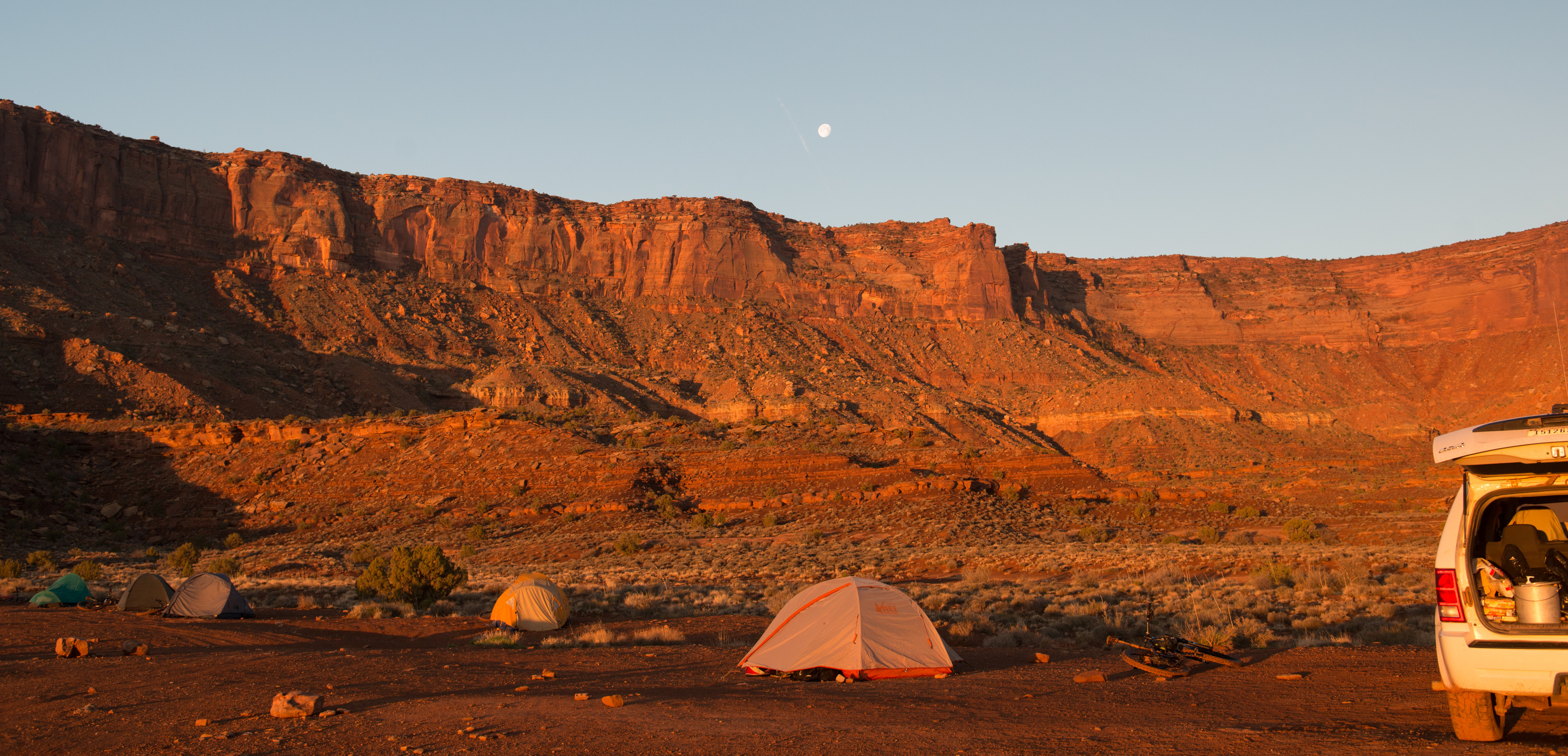 tents scattered around a flat area with tall orange cliffs and the moon in the distance