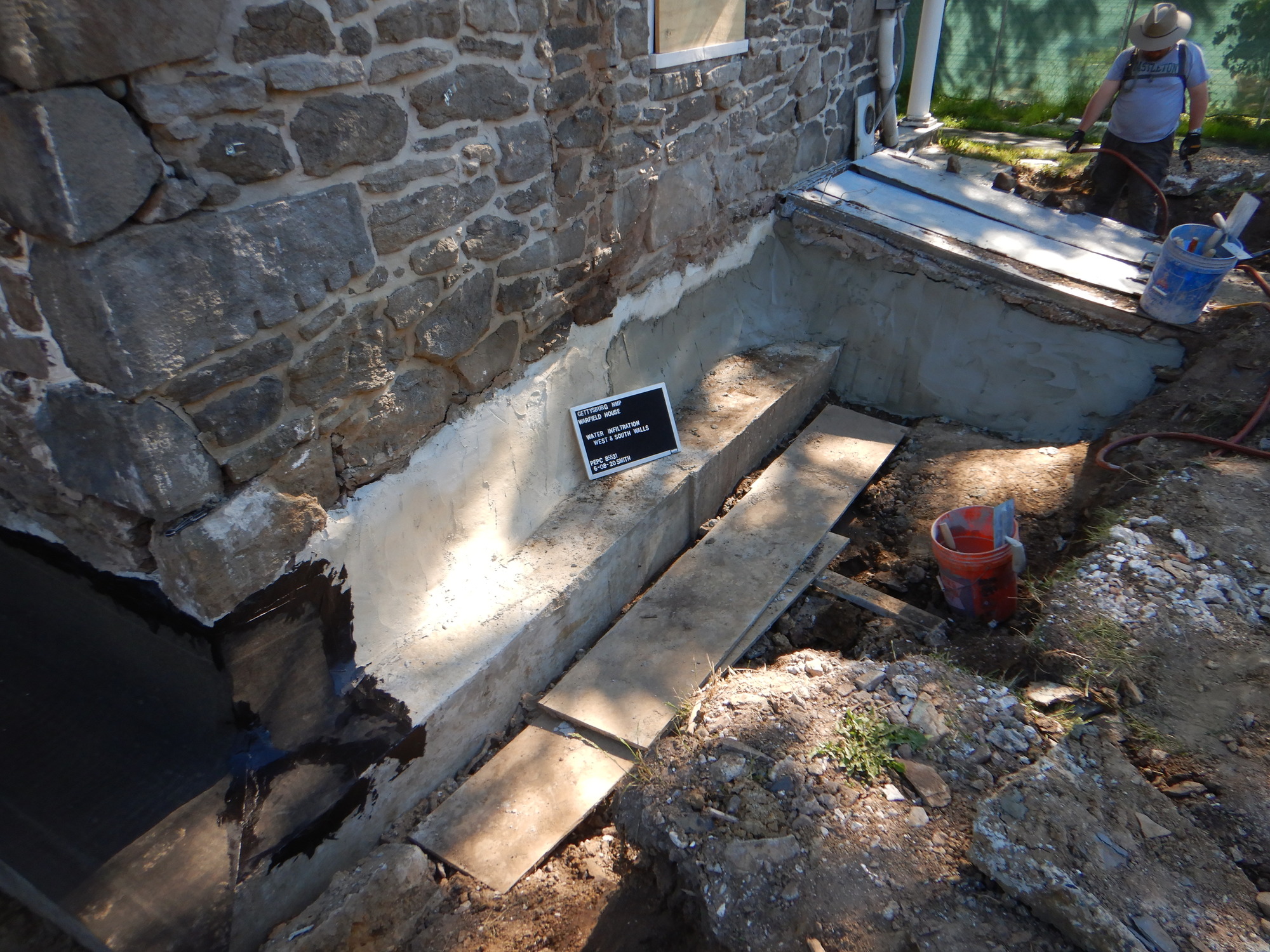 A NPS worker is seen in the background to the right. He is working on the rubber moisture barrier. The historic masonry foundation is repaired with the footer installation. Construction tools and wood boards are seen near the newly installed footer.