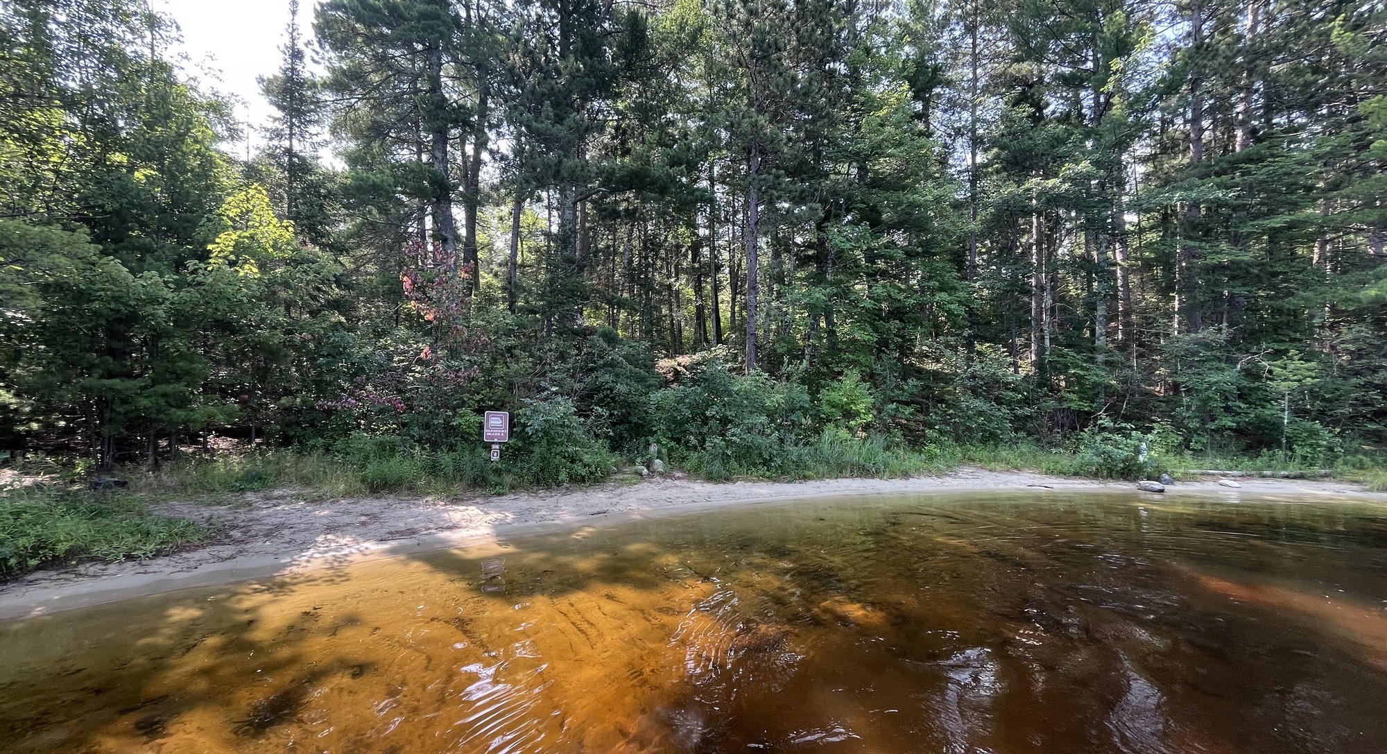 A calm, forested lakeshore with clear brown water, dense trees, and a small houseboat sign near the shoreline.