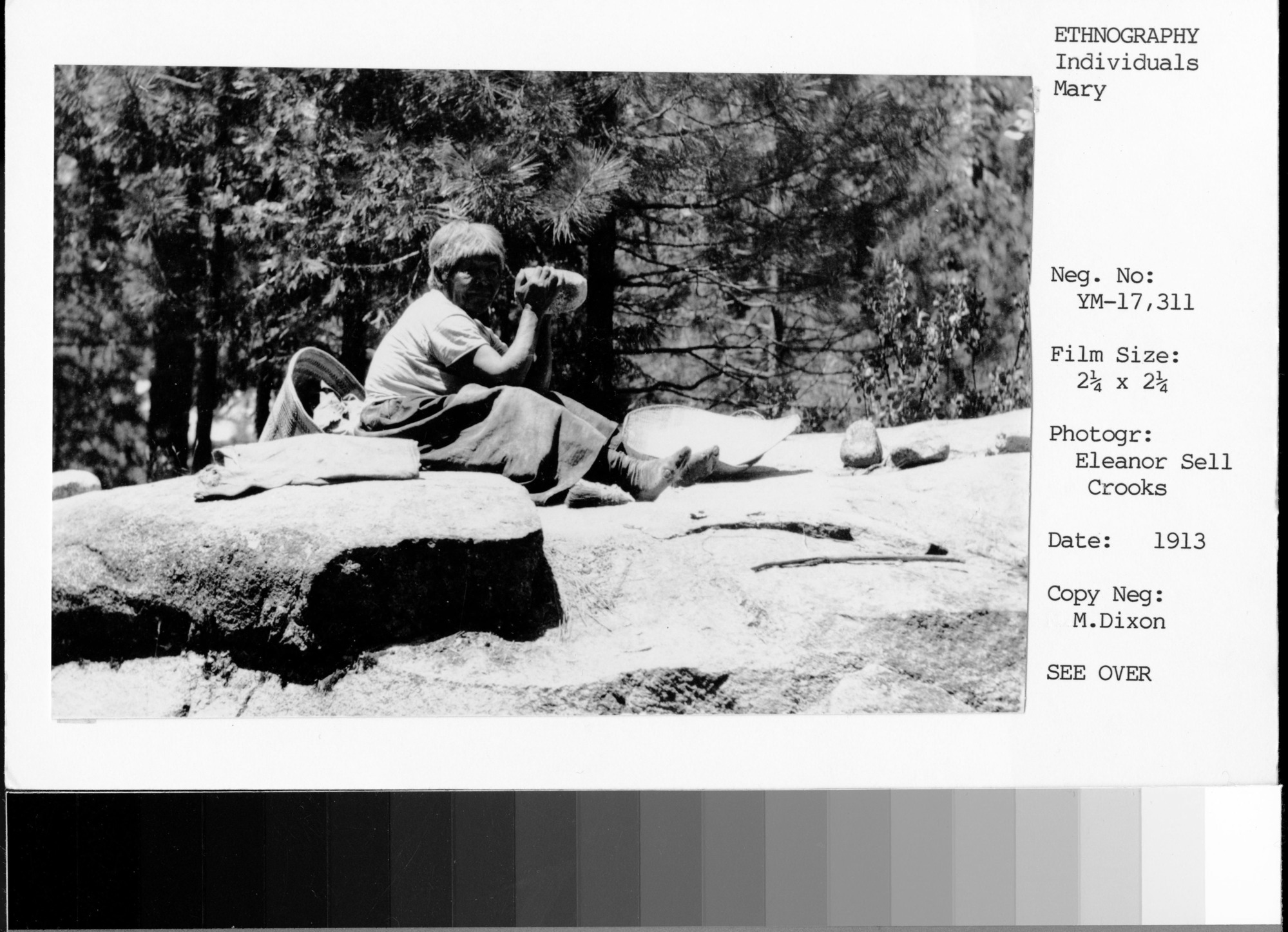Copy Neg: M. Dixon; Mary pounding acorn near Sentinel Creek, Yosemite Valley. An acorn flour sifting basket and a brush made of soaproot fibers are on the rock near Mary's feet, and a conical burden basket is behind her. Used in Traditions & Innovations, p. 18, fig. 29.