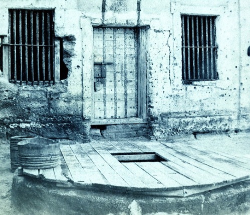 Black and white image of original door.  Bared windows and an old well in the foreground.