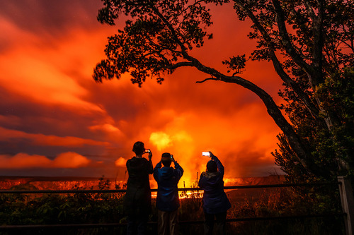 Three people use their mobile devices to photograph a glowing volcanic eruption in darkness. A tall tree is silhouetted and towers next to them. 