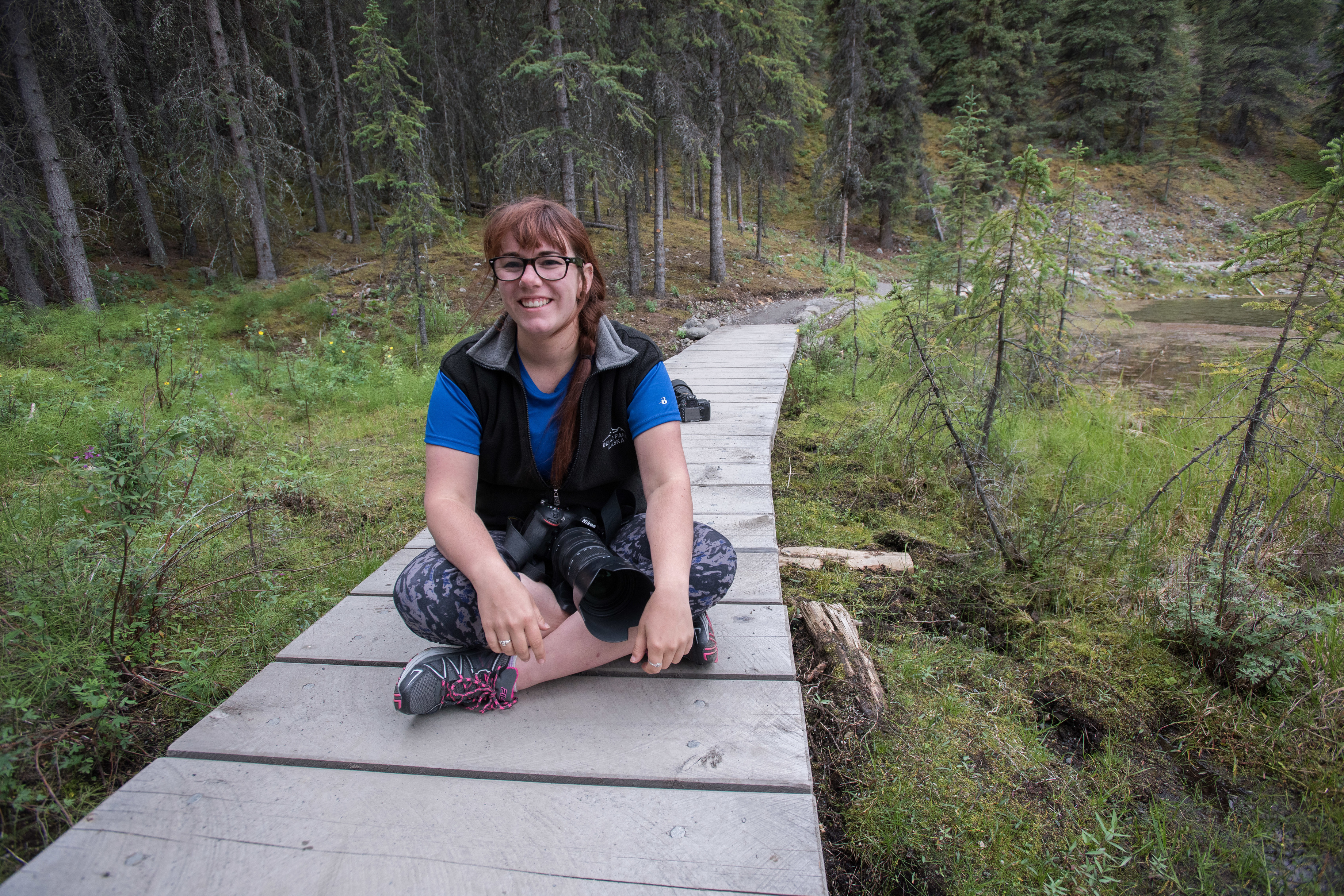 young woman with camera sitting on a boardwalk in a forest