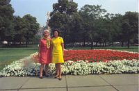 Two women in brightly-colored dresses pose on a sidewalk in front of a flower bed, with white-blooming flowers forming a border around red blooming flowers. 