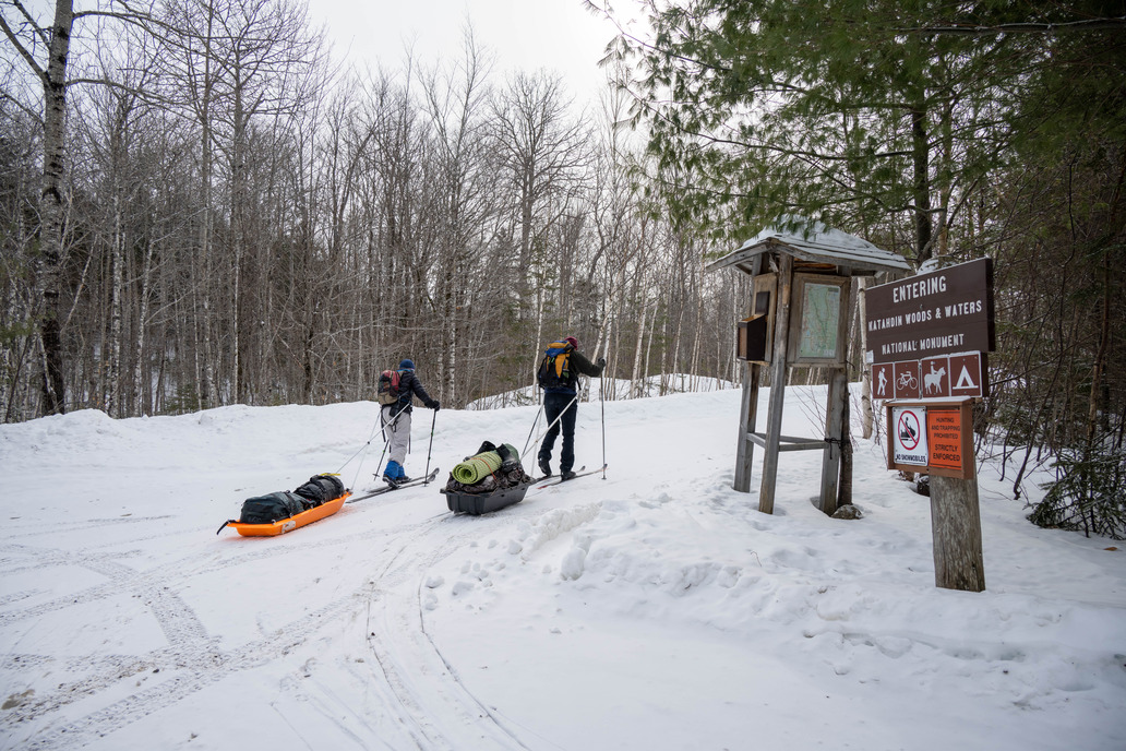 Two people on skis pull sleds behind them down a snow-covered trail. Trees without leaves and pine trees with needles sit in the background. Two brown wooden signs sit in the snow. 
