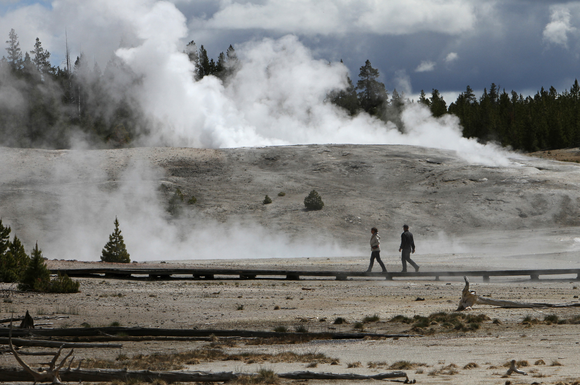 Two people walk from right to left on a boardwalk in a geyser basin with a steaming thermal feature in the background.
