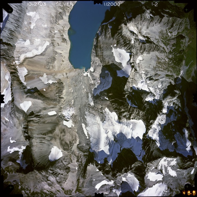 Color aerial photograph of glacier, snowfields, and alpine lake on mountain peak with forested valleys below.