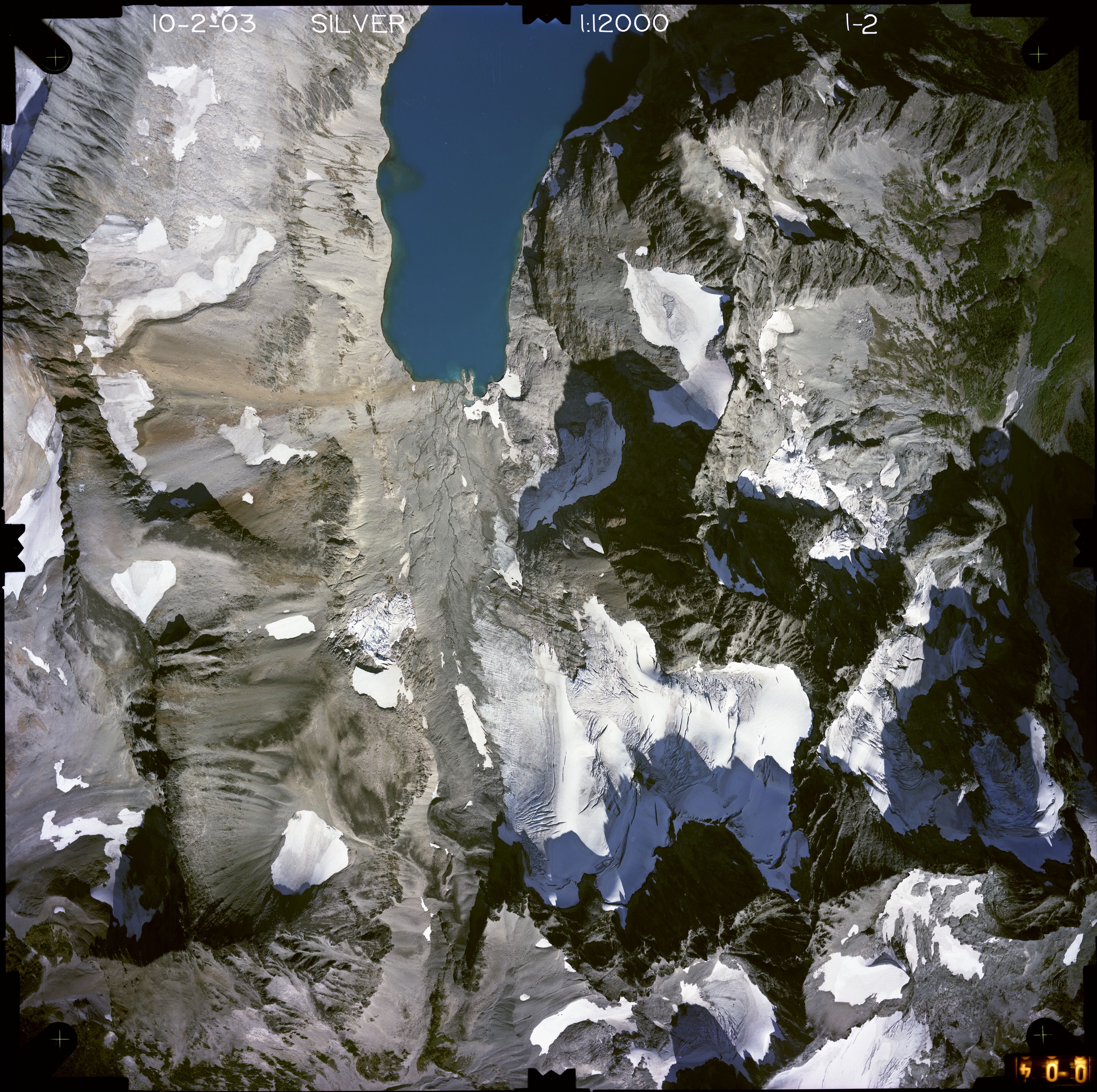 Color aerial photograph of glacier, snowfields, and alpine lake on mountain peak with forested valleys below.