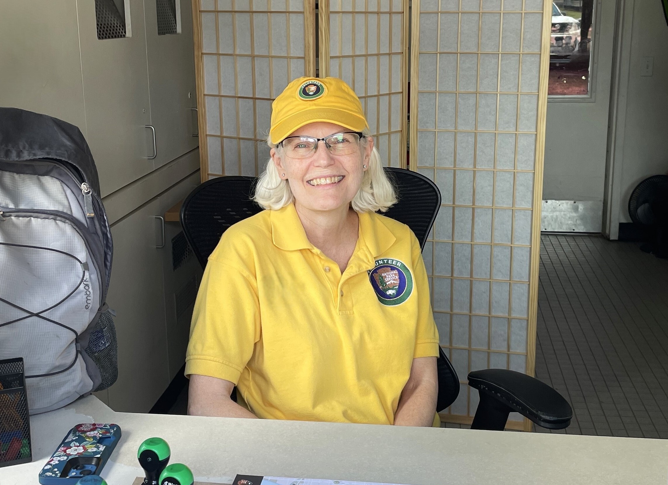 A woman wearing a yellow shirt, yellow hat, and glasses sits at a desk while smiling for a photograph.