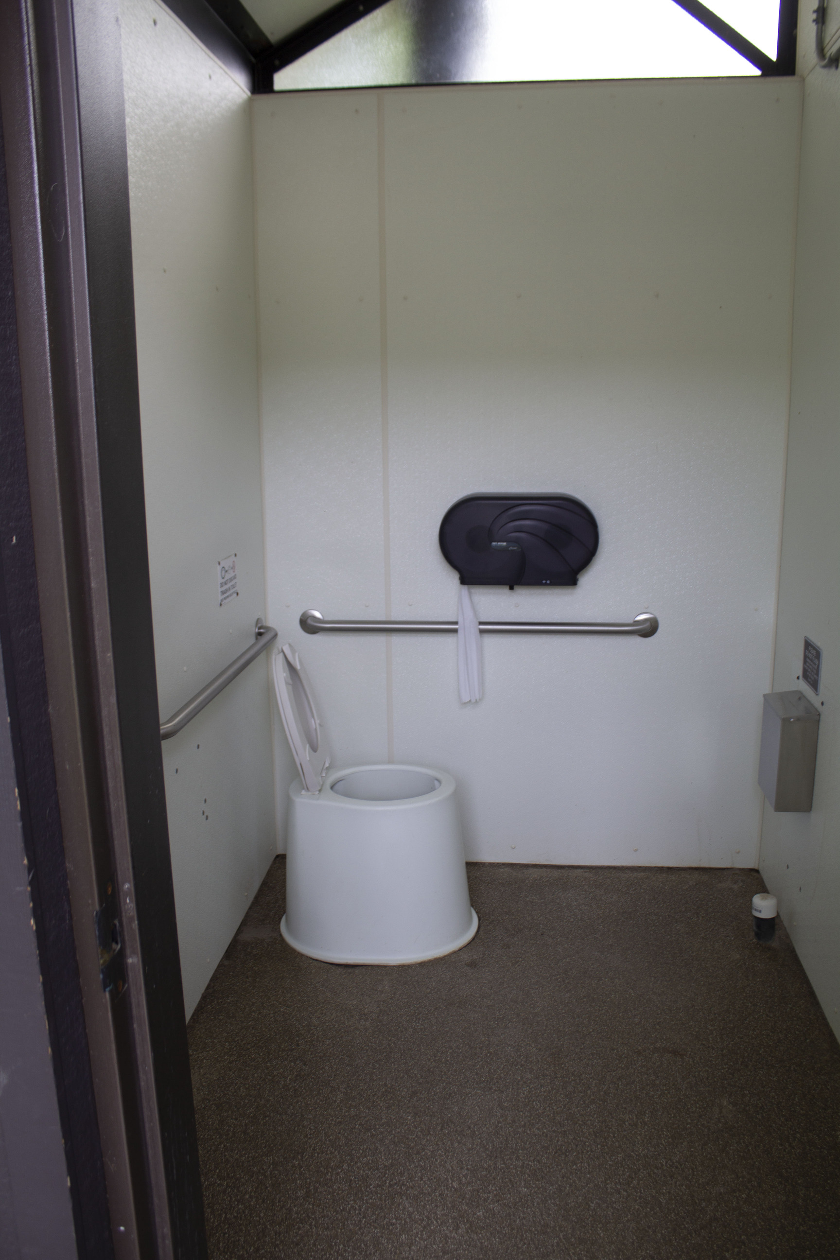 Toilet sits in the far left of a small room with a waste container mounted on the opposite wall.