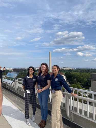 3 people stand near a railing, the Washington monument in the distance behind them.