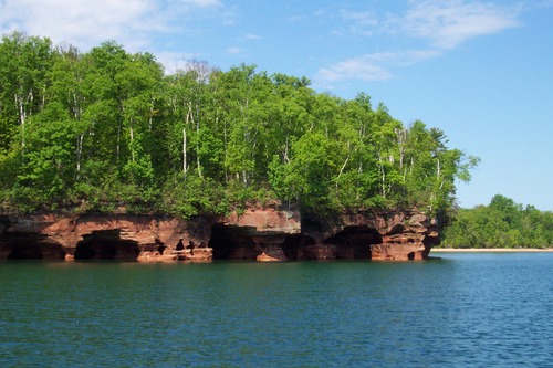 Red sandstone sea caves covered by trees on the side of a lake and a sandy beach in the distance.