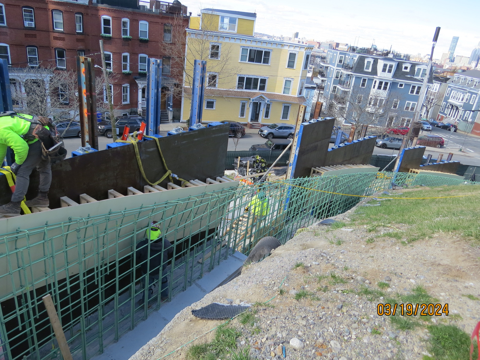 Green rebar is arranged in the shape of a wall lining the bottom of a hill. On the side away from the hill are metal barriers. 