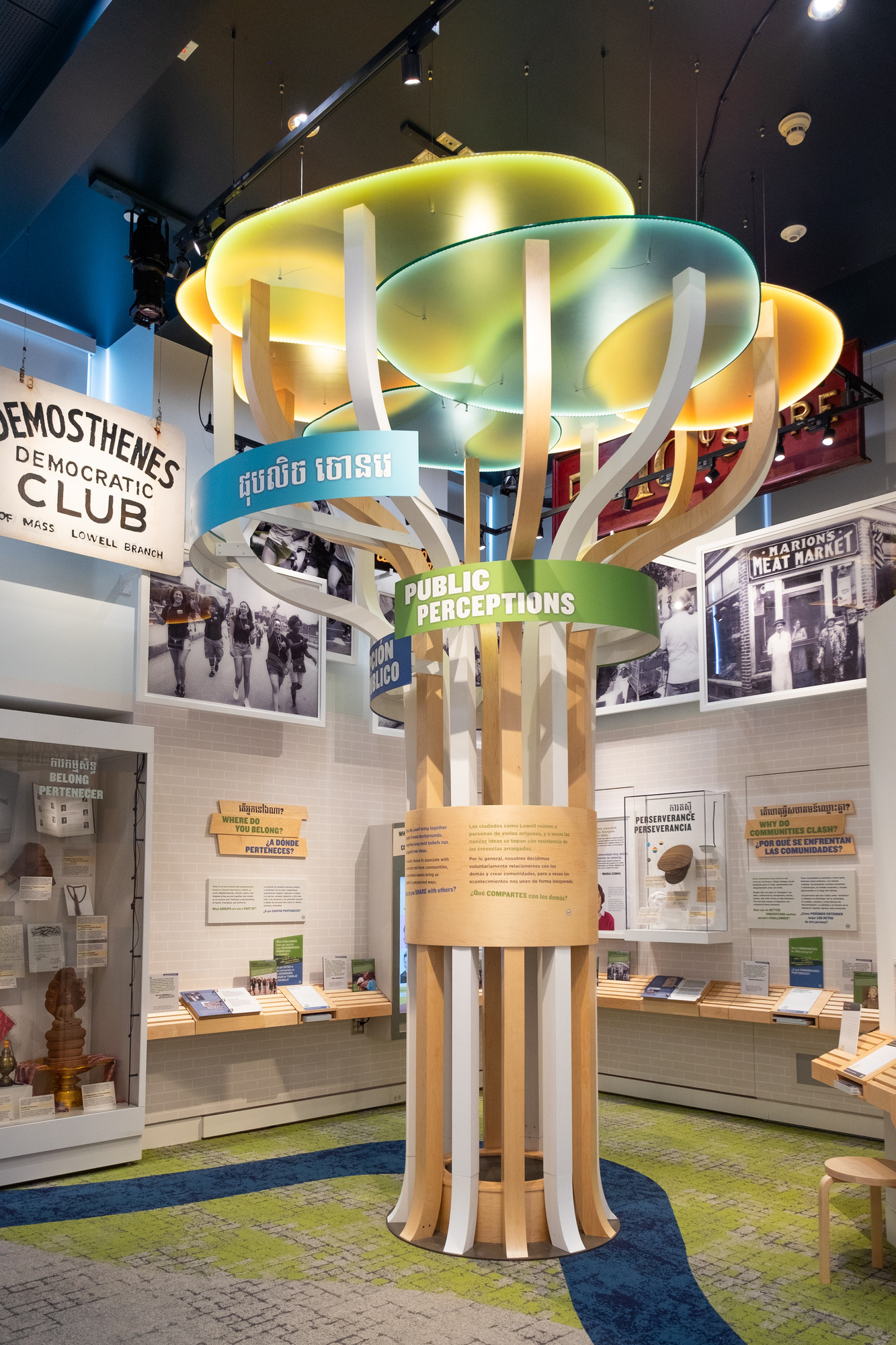 In the background hanging from the ceiling are historic and photos and shops signs. Centered in frame is a large artistic interpretation of a tree with the words "Public Perceptions" written in English, Spanish and Khmer. The tree's truck and branches are made from wooded beam, and the leaves are lit colorful panes.