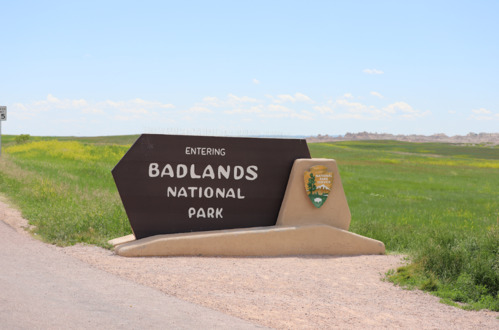a large brown sign with a national parks service logo reads "entering badlands national park" with green prairie behind it and badlands buttes in the far distance