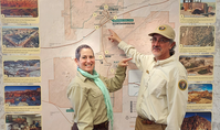 Woman and man in uniform standing in front of a map with photographs.