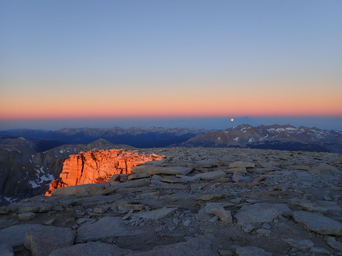 Orange sunlight hits a stone wall, while the moon sets in the distance
