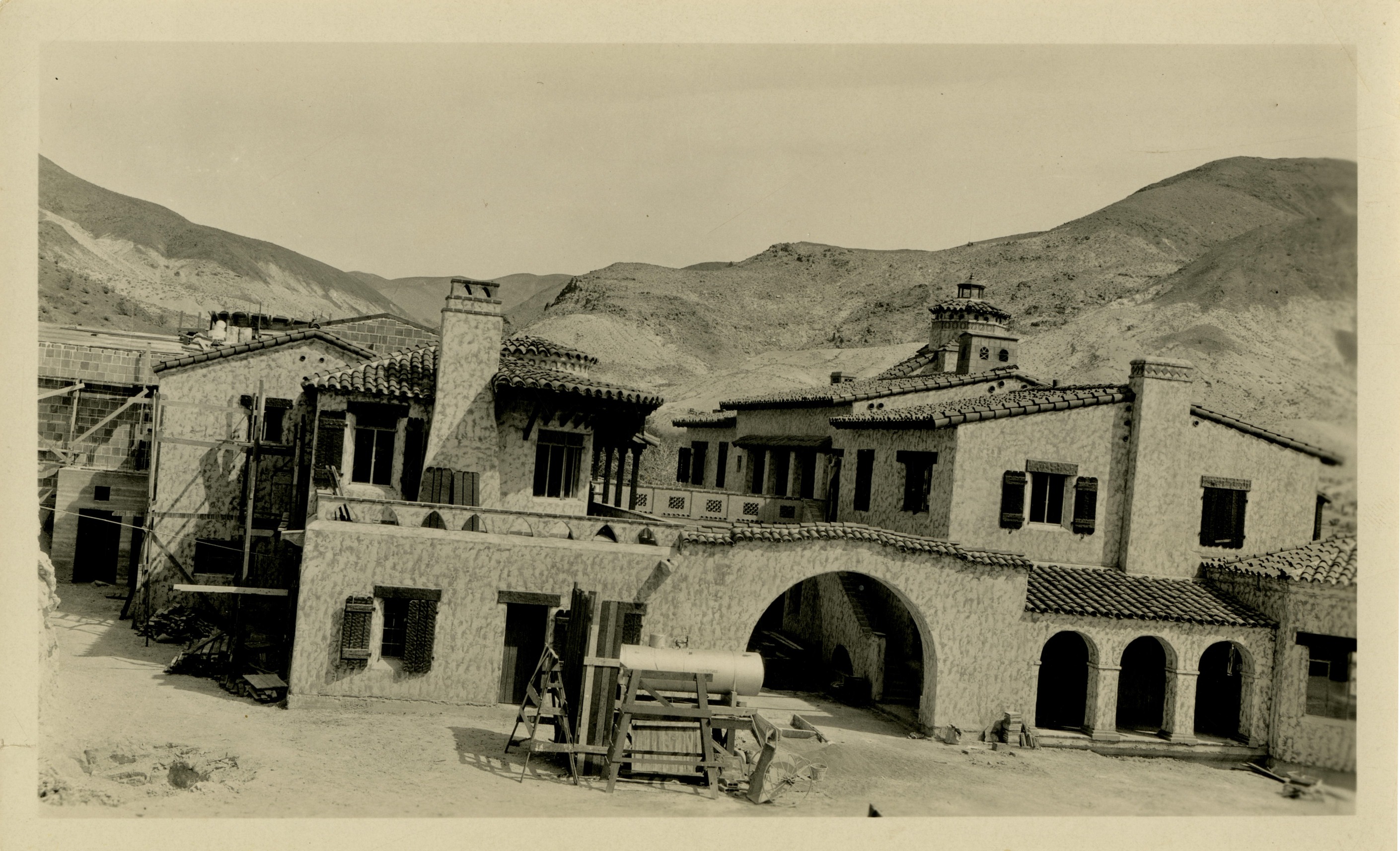 This is an historic black and white photograph from the Scotty's Castle Historic Photograph Collection, Death Valley National Park of a large, complex, Spanish-style structure newly constructed with some on-going construction. Scotty’s Castle Main House with octagon-shaped room on left, flying buttress front porch towards right, square Observation tower on right.  Scaffold, ladders scattered around foreground. Other structures in distance on right. Desert hillsides in background.