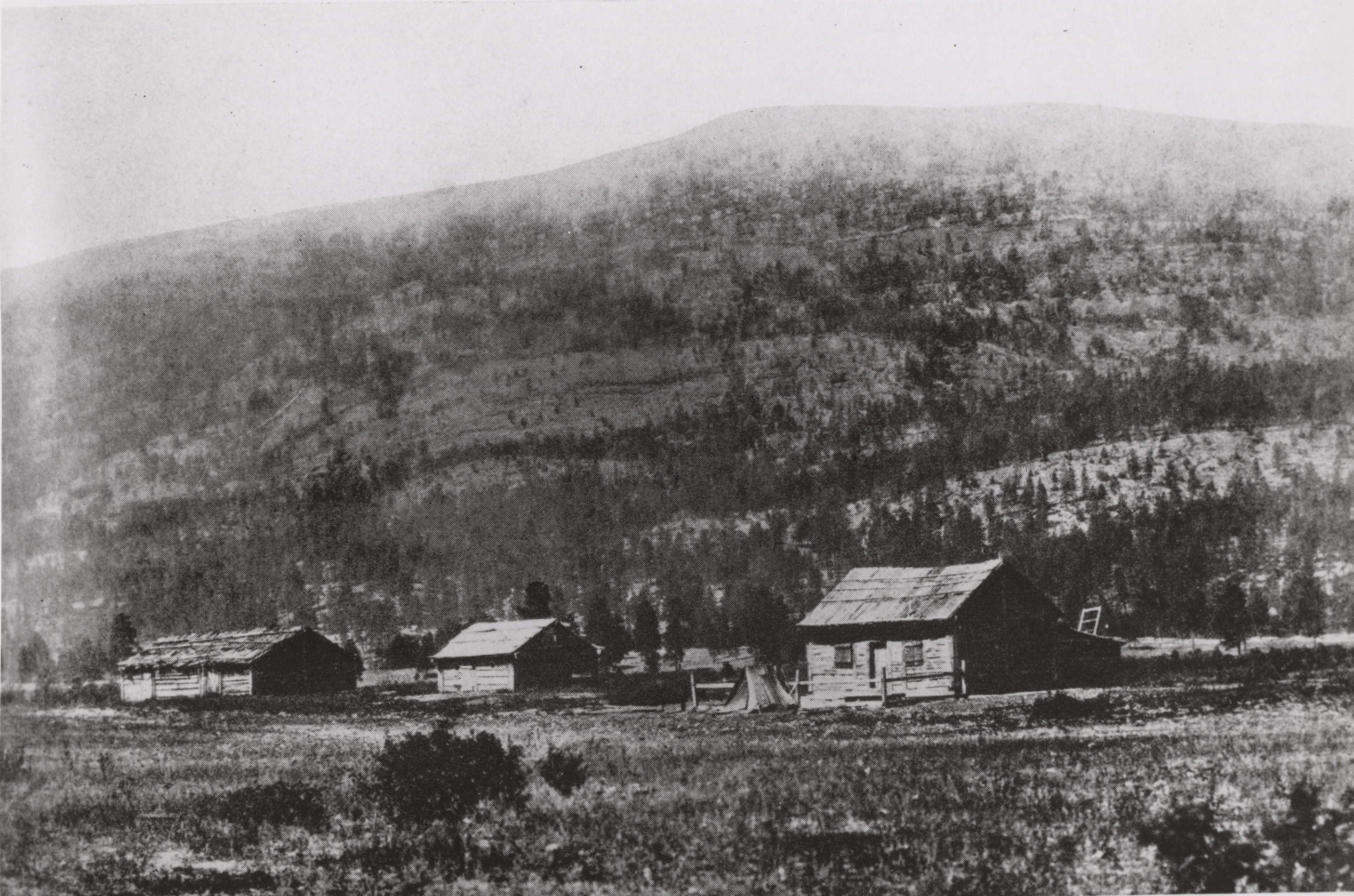 Black and white photograph of three wooden buildings at the base of a forested hill