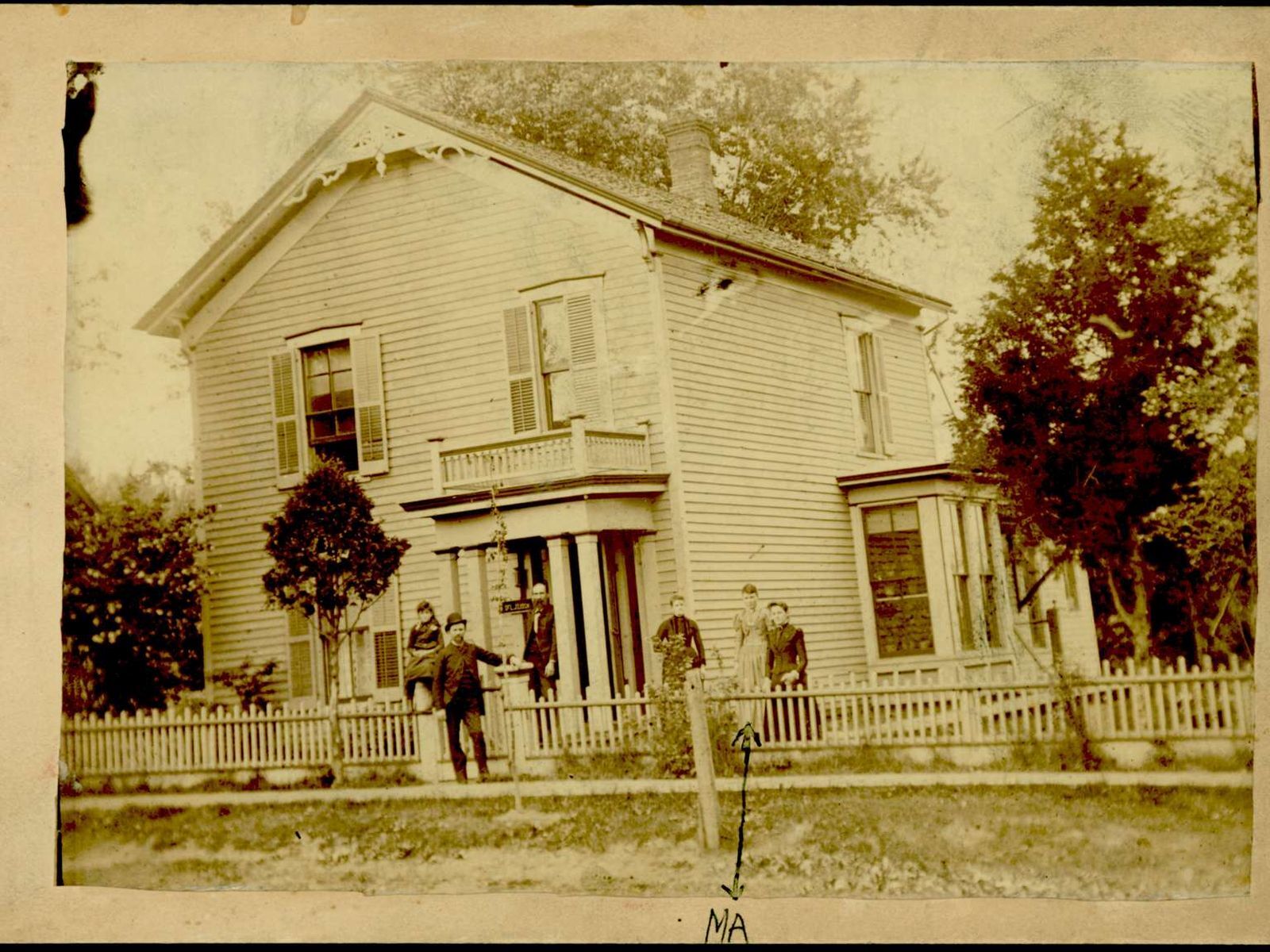 A family poses in front of a two story house, which has a white picket fence and a tree in front.
