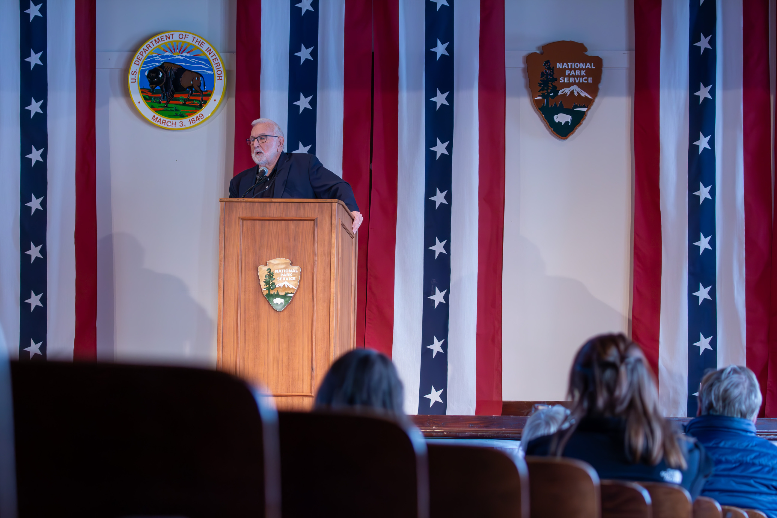 Dr. Stanly Godbold, Jr. standing on stage behind podium. Red, white, and blue flag banners, Department of the Interior symbol, and National Park Service arrowhead in background.
