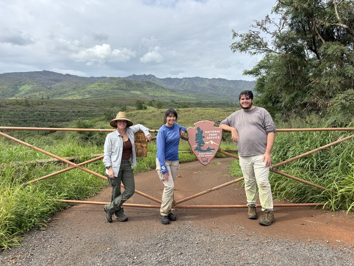 Three people are standing and smiling in front of a gate that has the NPS arrowhead on it.