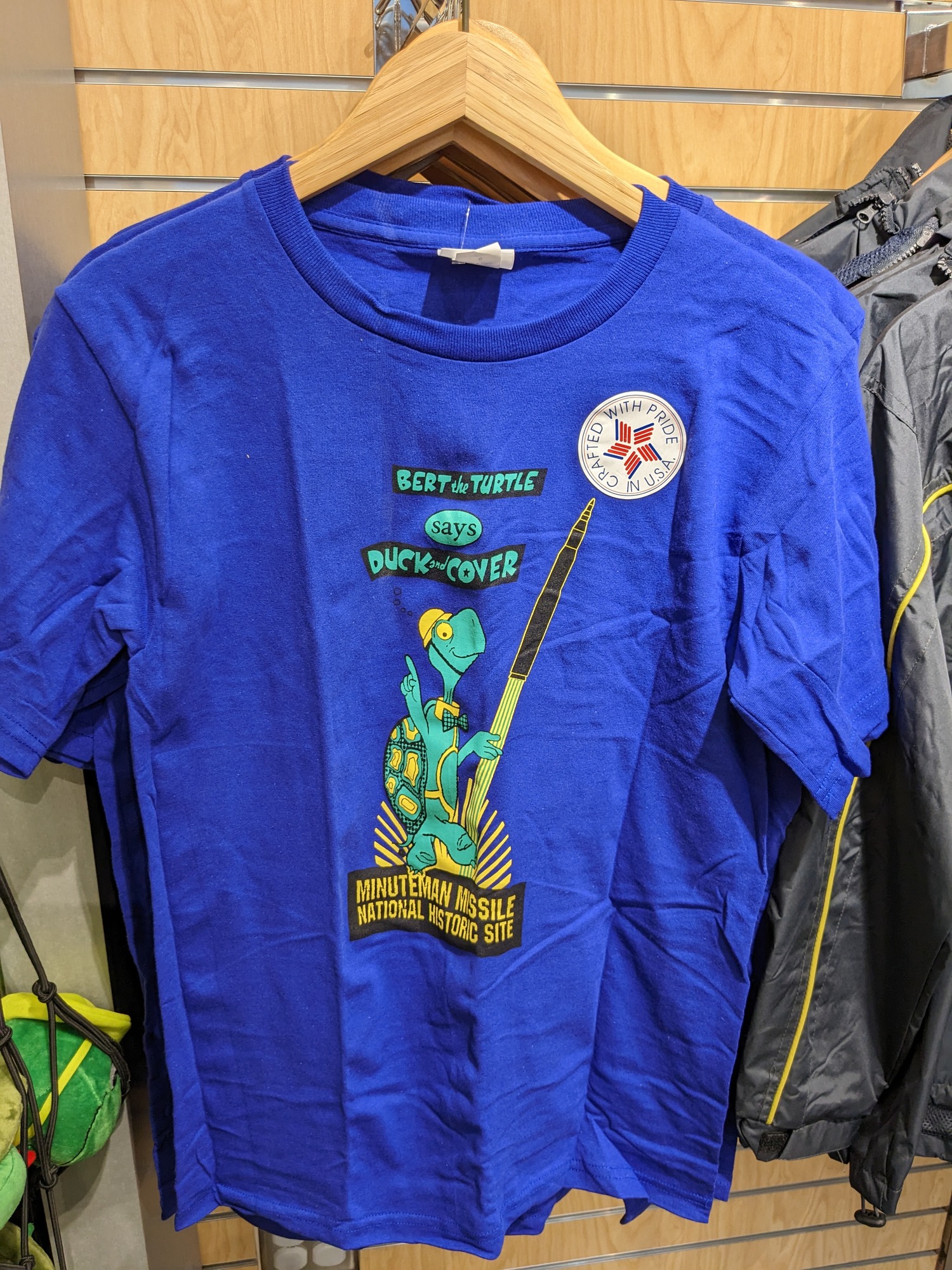 Blue t-shirt. Words in green "Bert the turtle says duck and cover". A green turtle with yellow helmet and a black missile. Words in yellow against a black background "Minuteman Missile National historic site" 