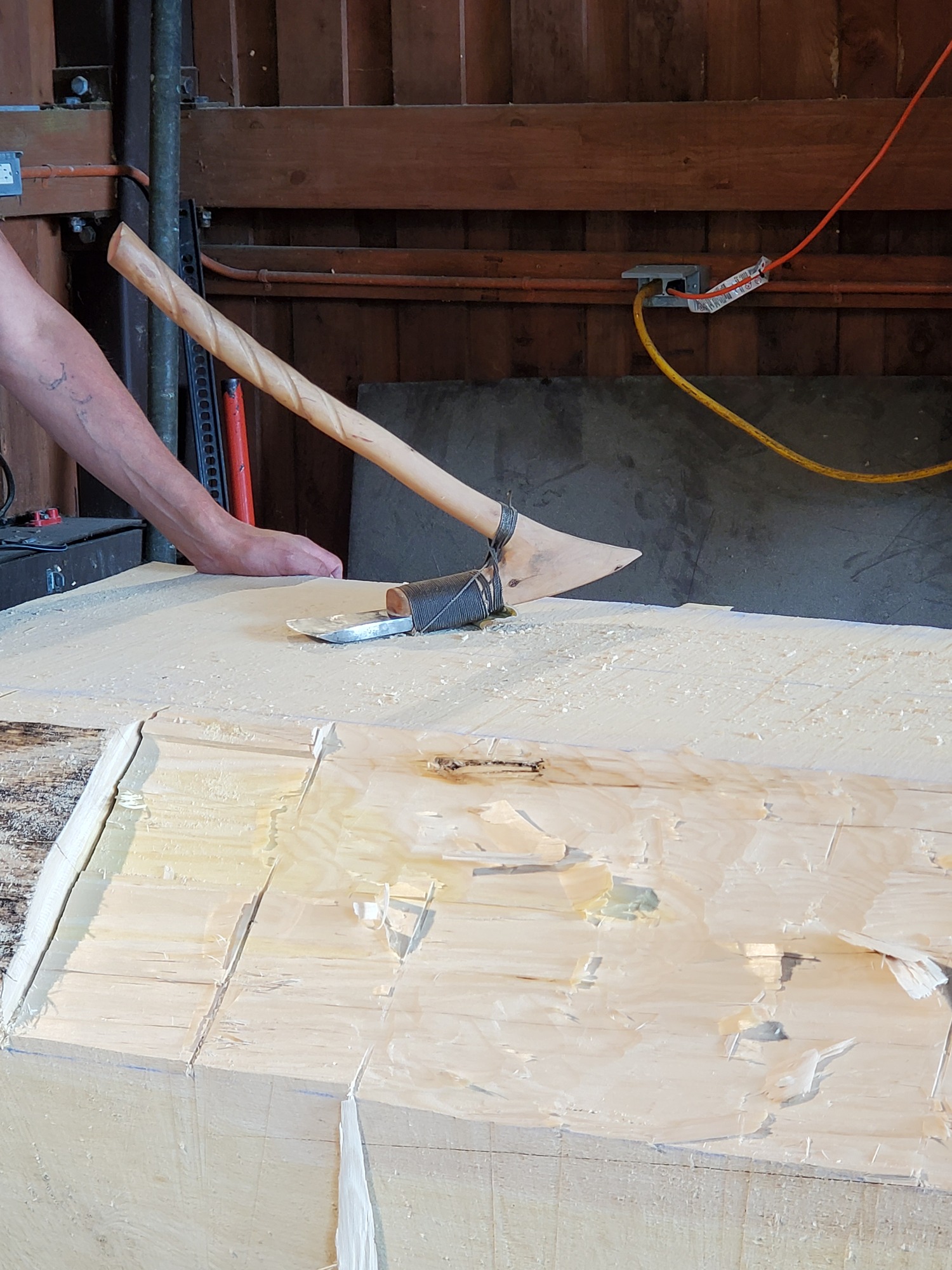 An Elbow Adze sits on top of a flat wooden surface. The carver's arm is visible on the left. 