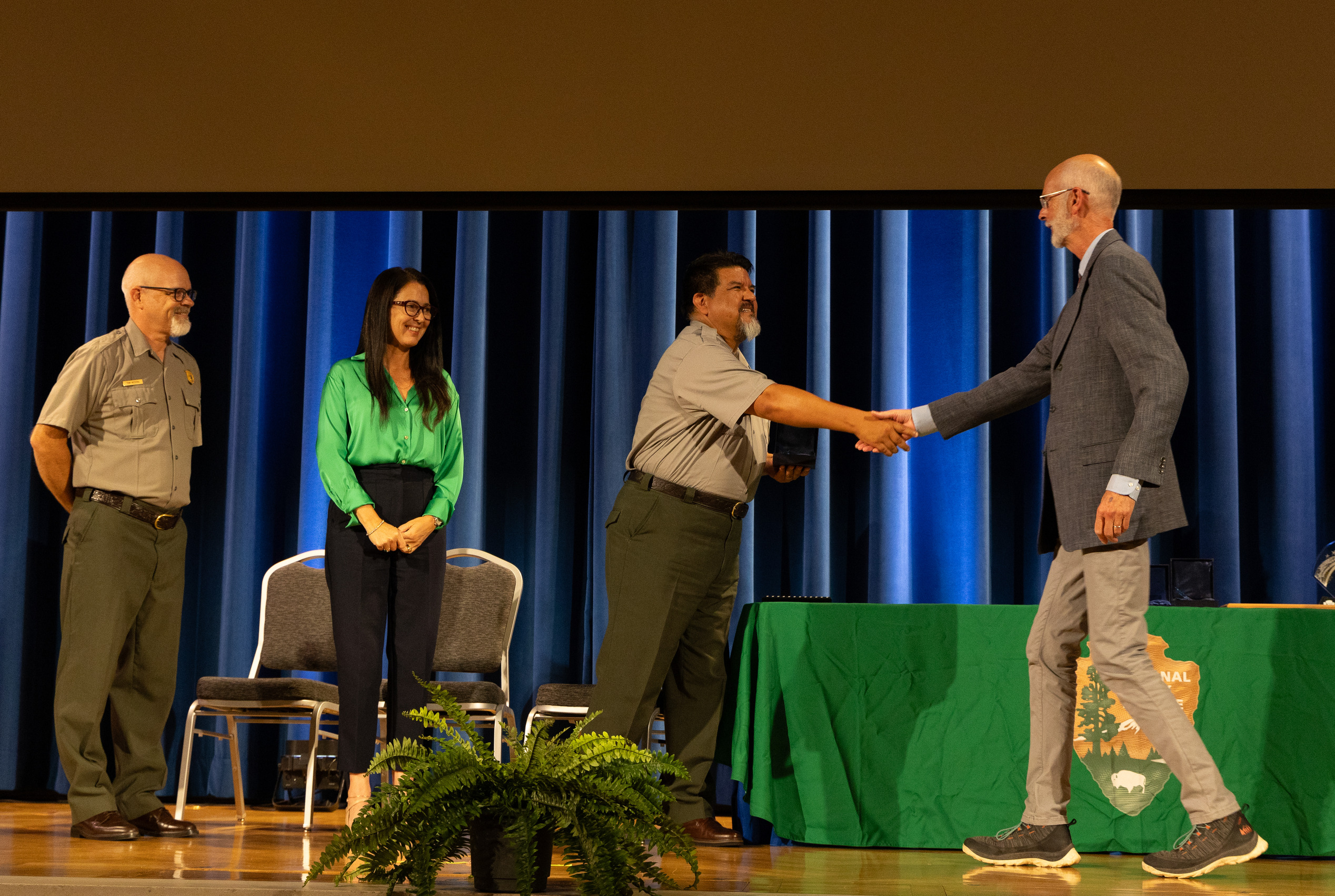 Two men shake hands on a stage during an awards ceremony. Two other people watch and smile at left.